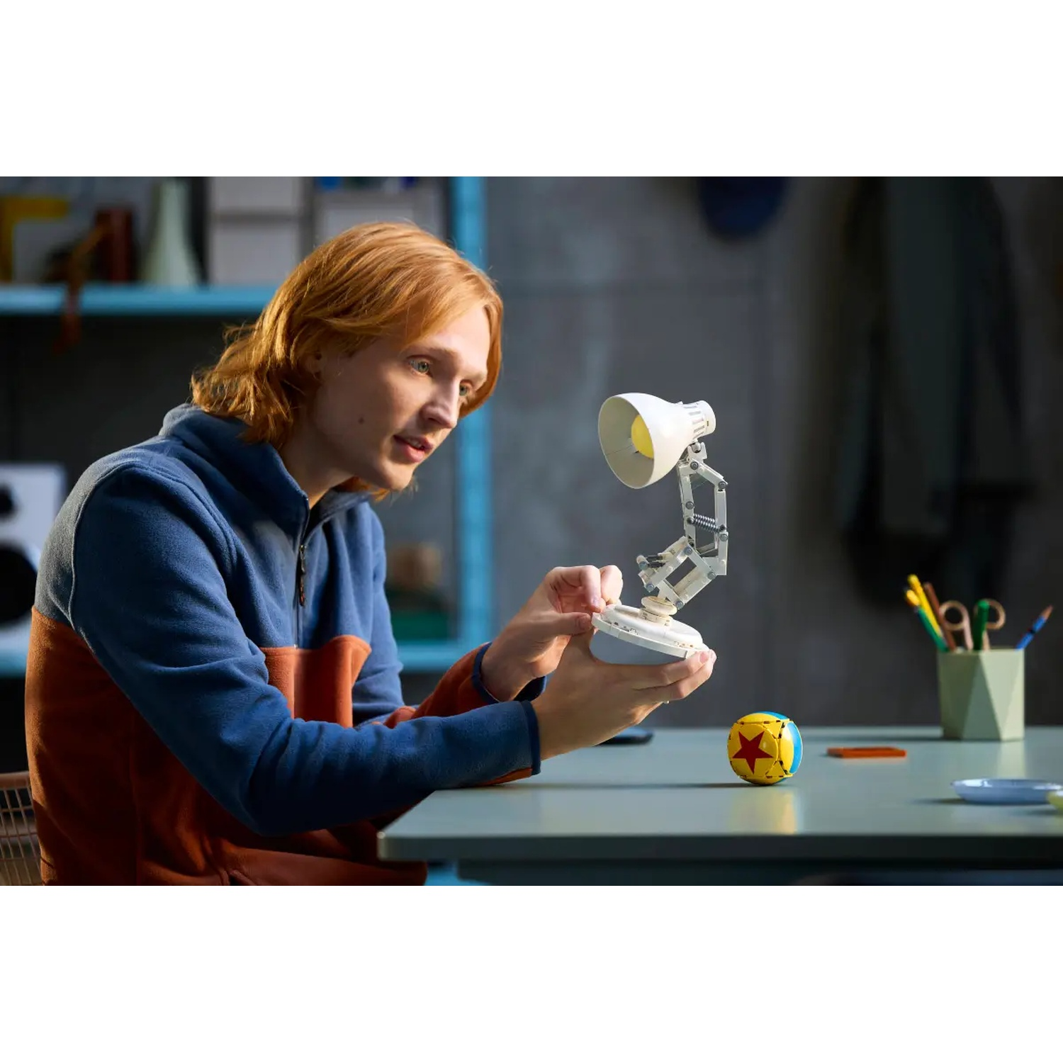 A person with red hair is assembling a white lamp. A yellow ball with a red star sits on the table. The background is blurred.