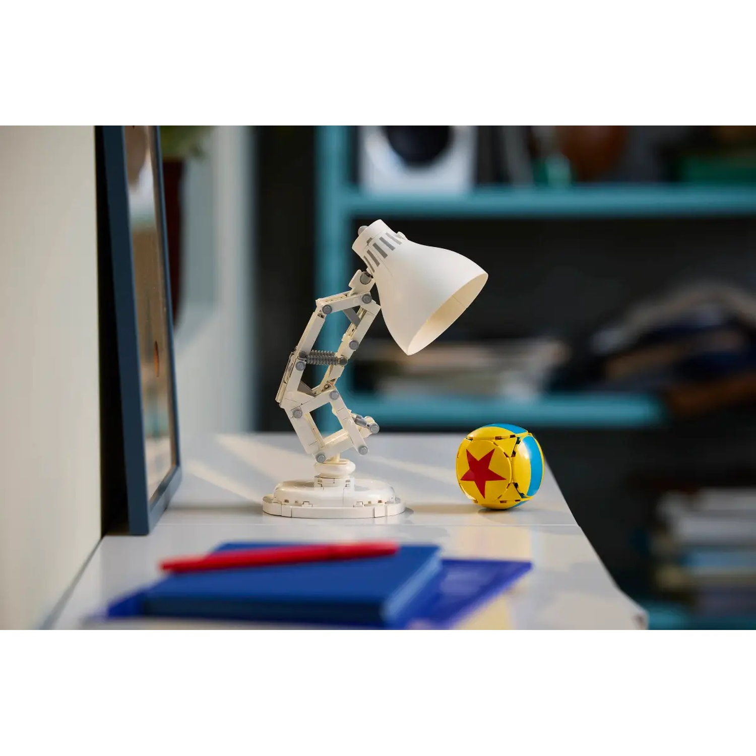 A white desk lamp and a Pixar ball are on a white surface. A blue notebook and a red pen are visible. A blurred background.
