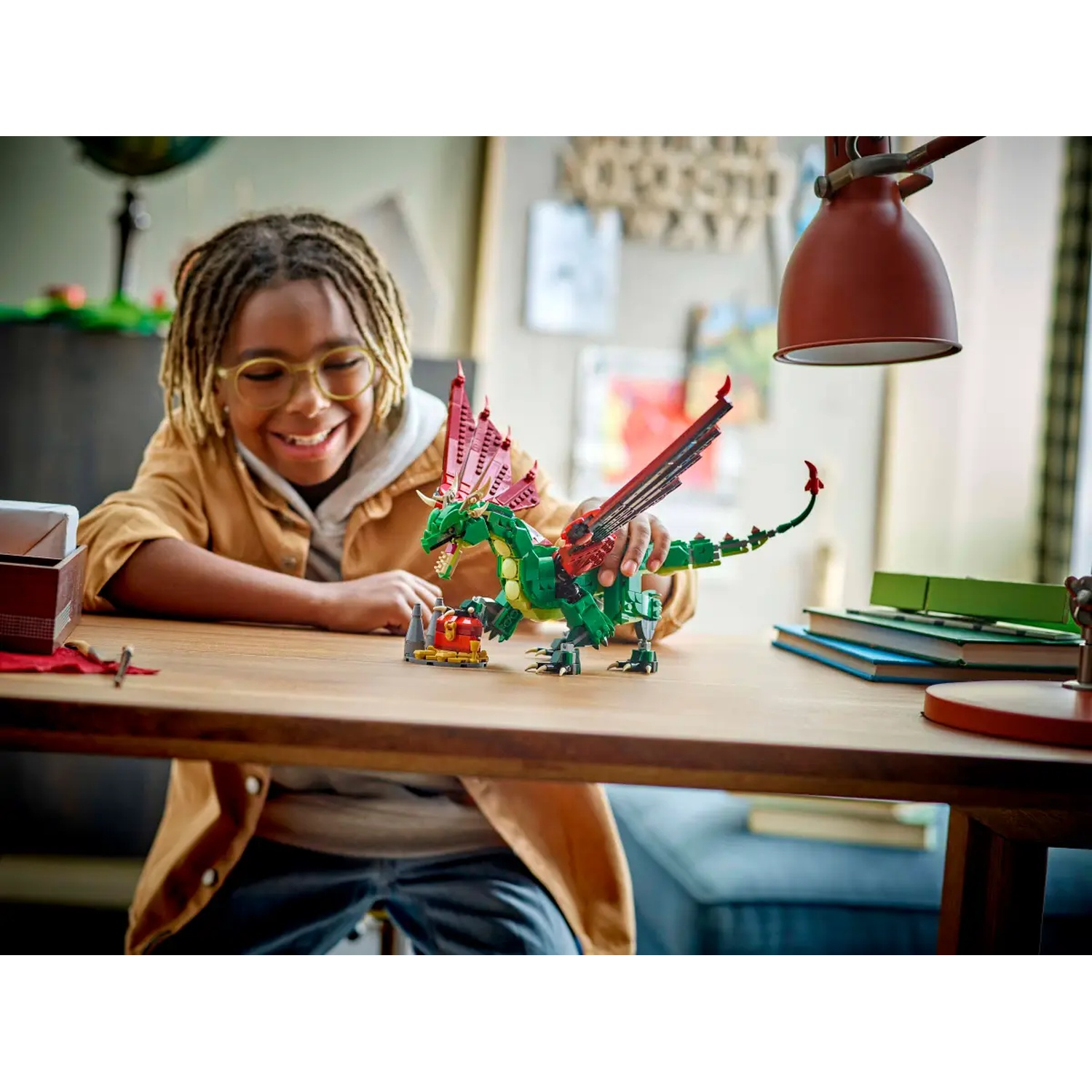 A smiling boy with glasses plays with a Lego dragon on a wooden table. A red lamp and books are in the background.