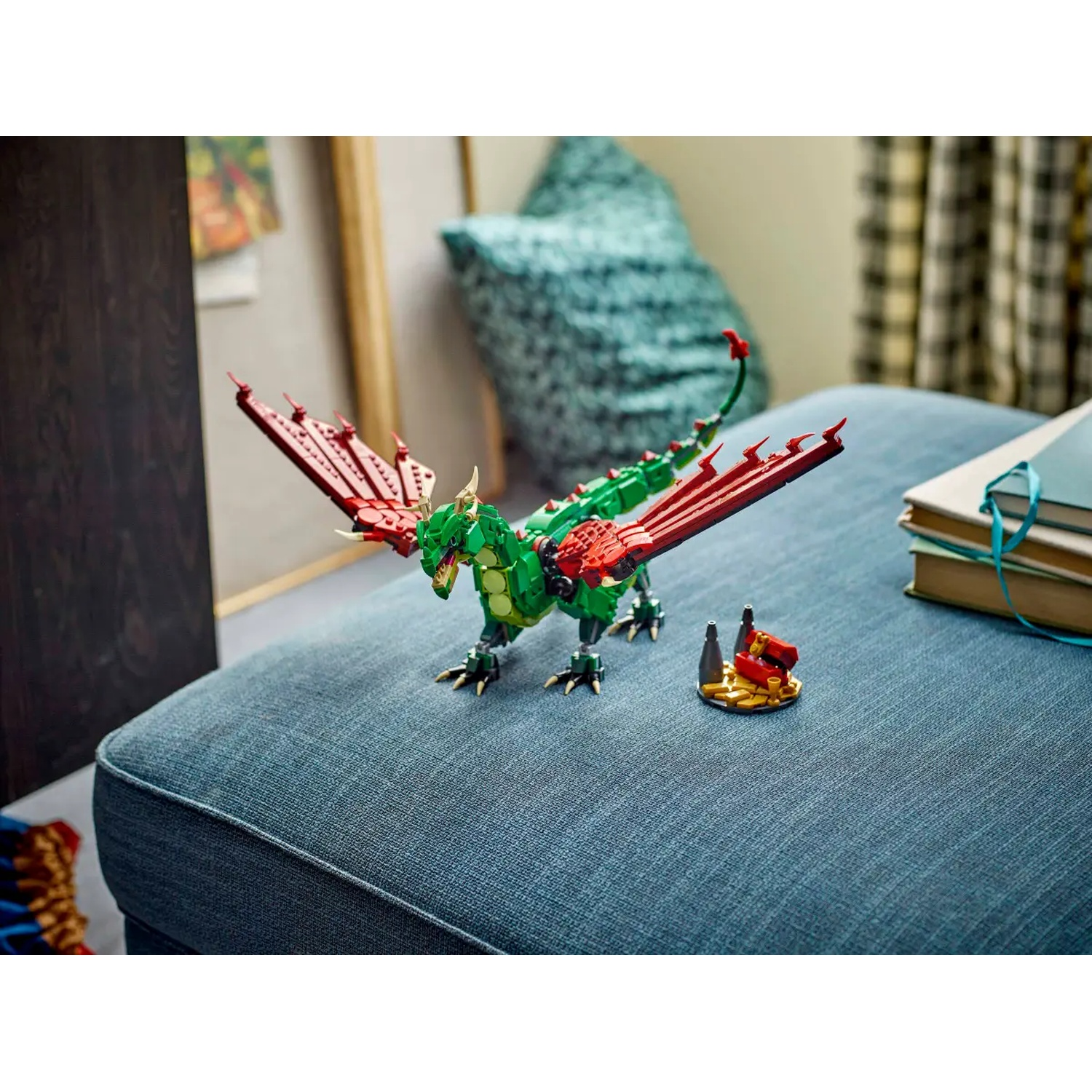 A Lego dragon with red wings, green body, and yellow claws sits on a blue ottoman. A small Lego structure is nearby.