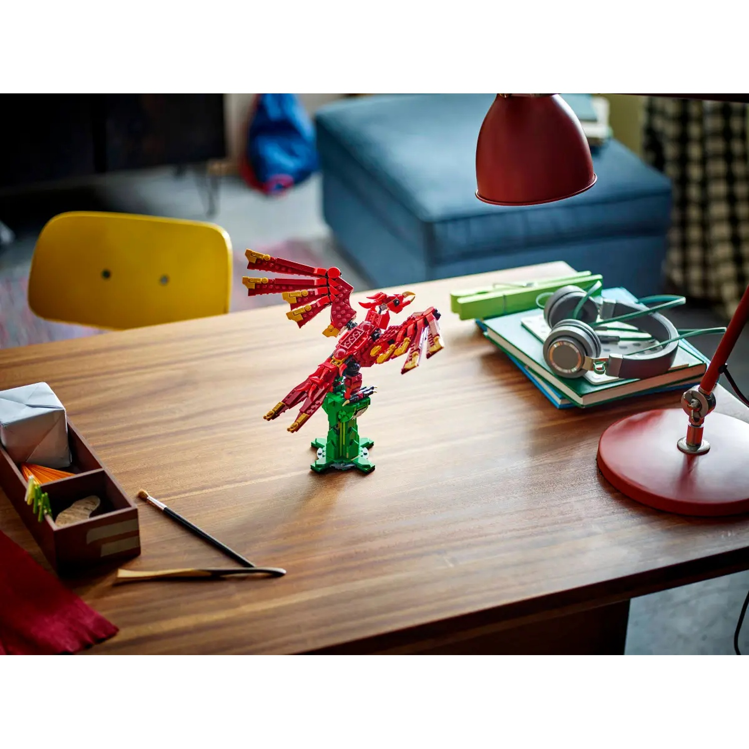 A Lego phoenix model on a wooden table, with a red lamp, books, and headphones. A yellow chair and blue couch in the background.