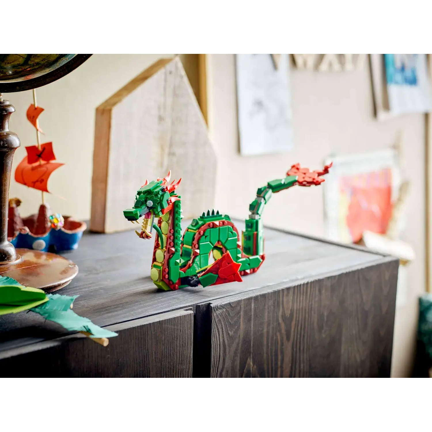 A Lego dragon, green and red, sits on a wooden cabinet. A globe and paper boats are also on the cabinet.
