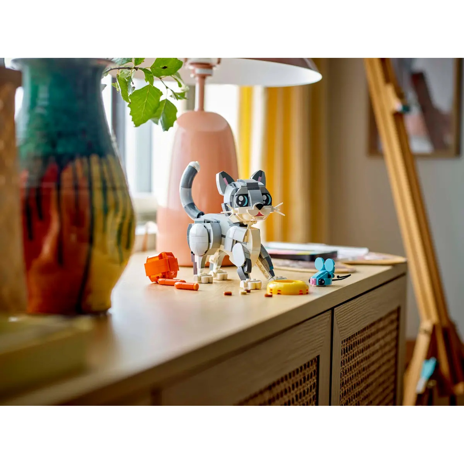 A Lego cat, orange and yellow objects, and a blue mouse sit on a wooden surface, with lamps and a vase in the background.