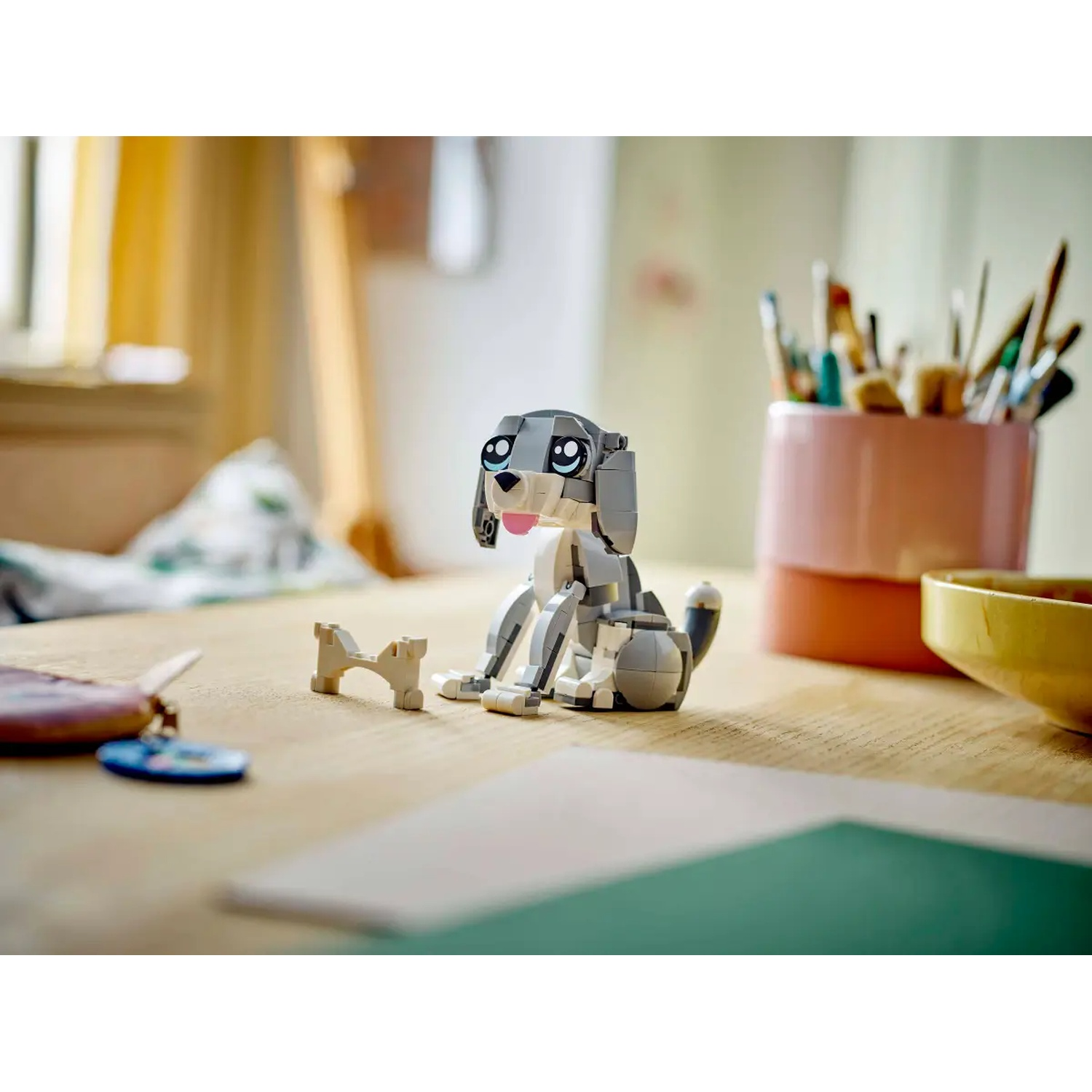 A Lego dog, gray and white, sits on a wooden table next to a bone, a key ring, and a bowl. Paintbrushes in background.