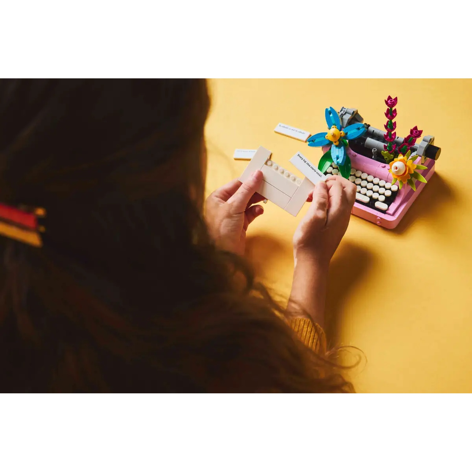 Een persoon die een roze Lego-typemachine met bloemversieringen op een gele ondergrond in elkaar zet, met witte Lego-stukken.