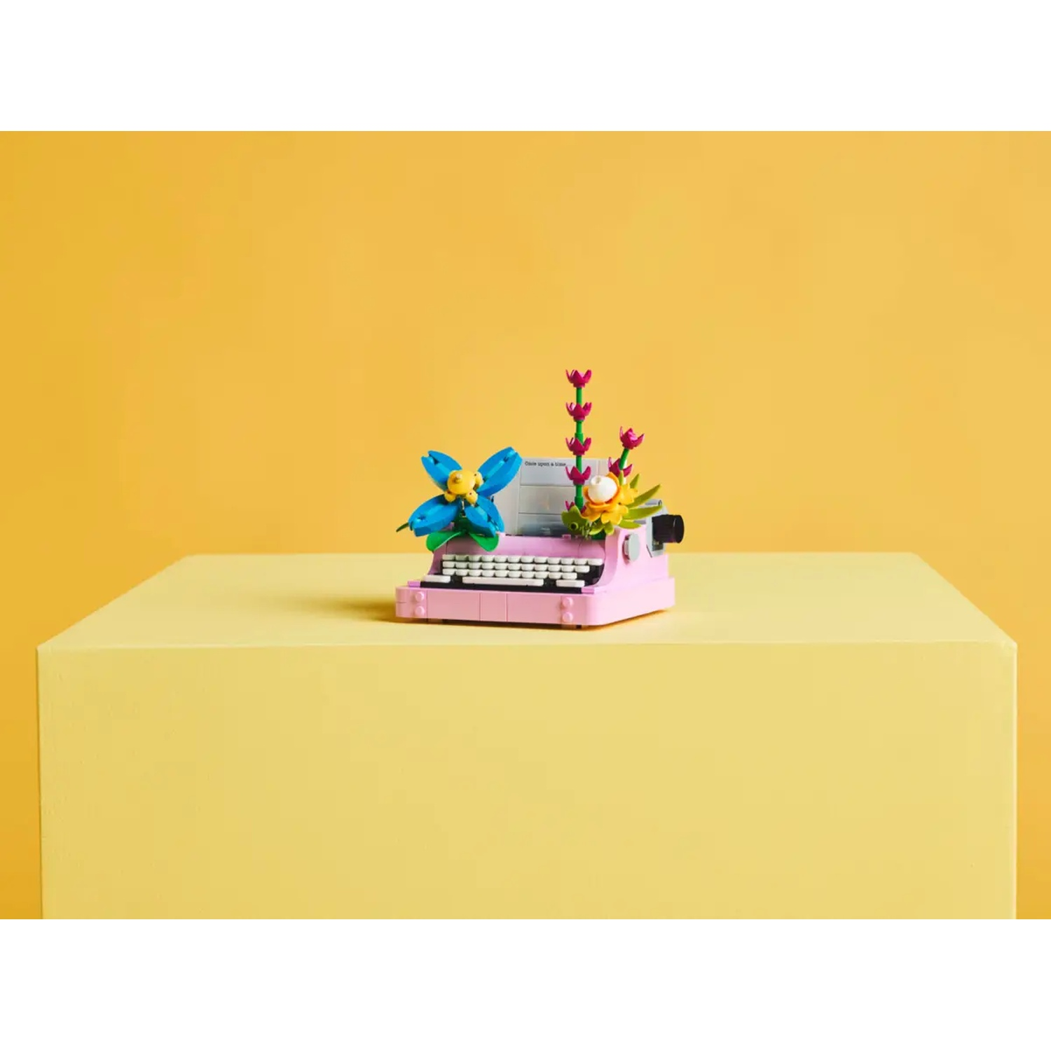 Een roze Lego-typemachine met blauwe en gele bloemen erop, op een gele blok. Achtergrond is geel.