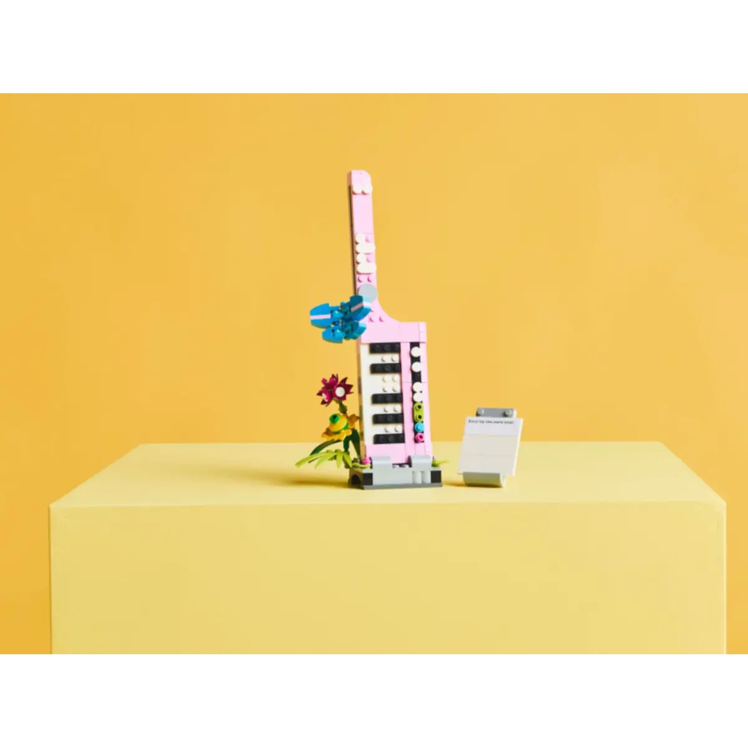 Een roze Lego-structuur met een blauwe vleugel, bloemen en een klein bord, op een geel oppervlak tegen een gele achtergrond.