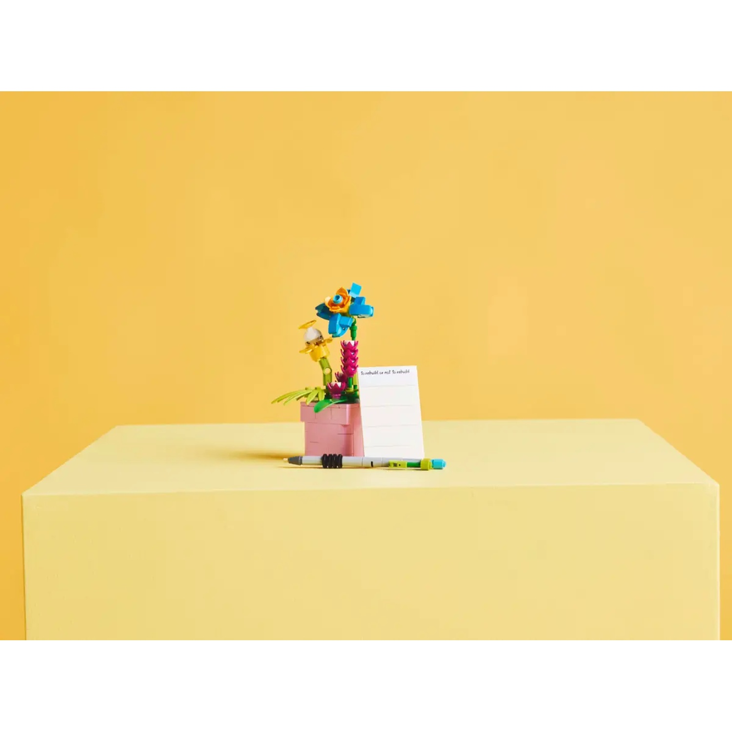 Een Lego bloemstuk staat op een geel oppervlak tegen een gele achtergrond. Een pen ligt ernaast.