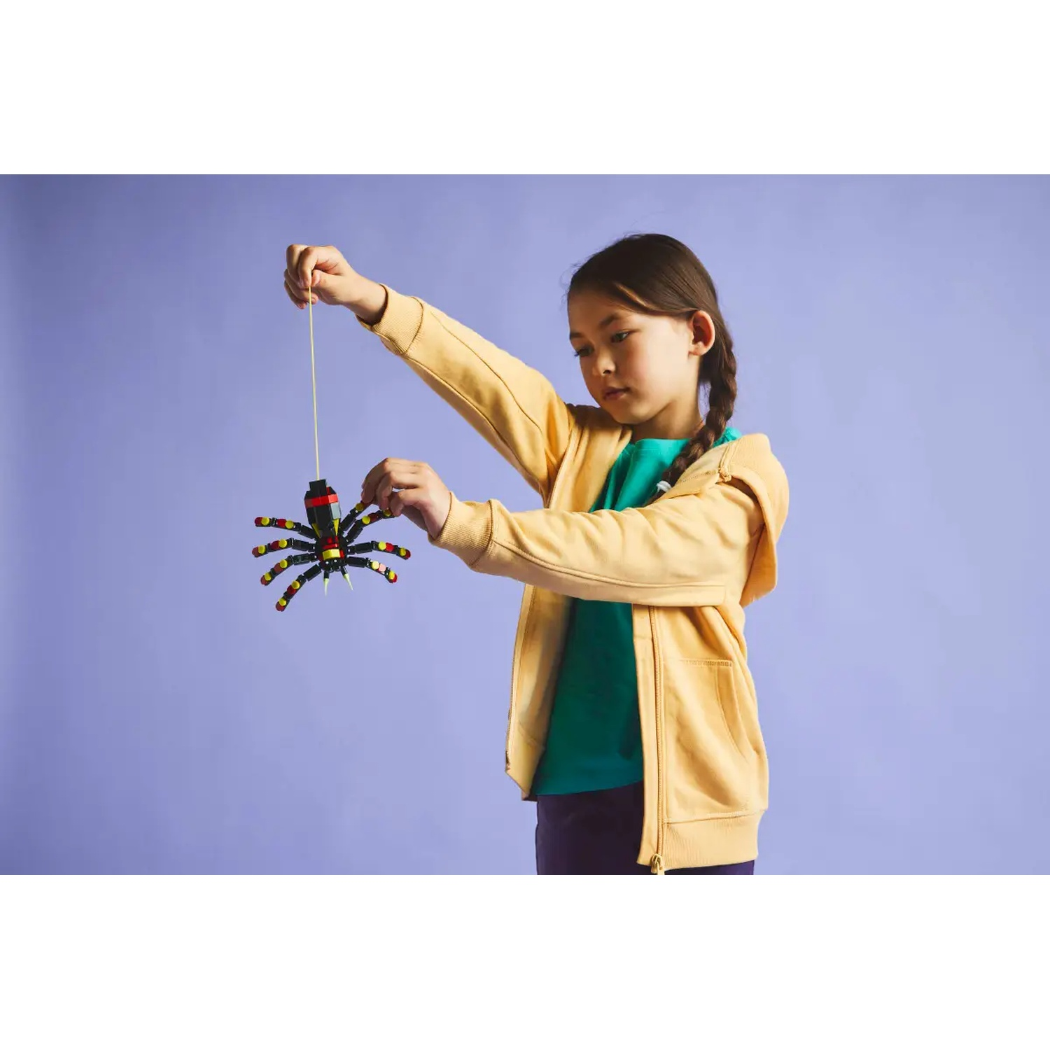 A girl in a yellow hoodie holds a toy spider. The spider is black with yellow and red details, suspended by a string.