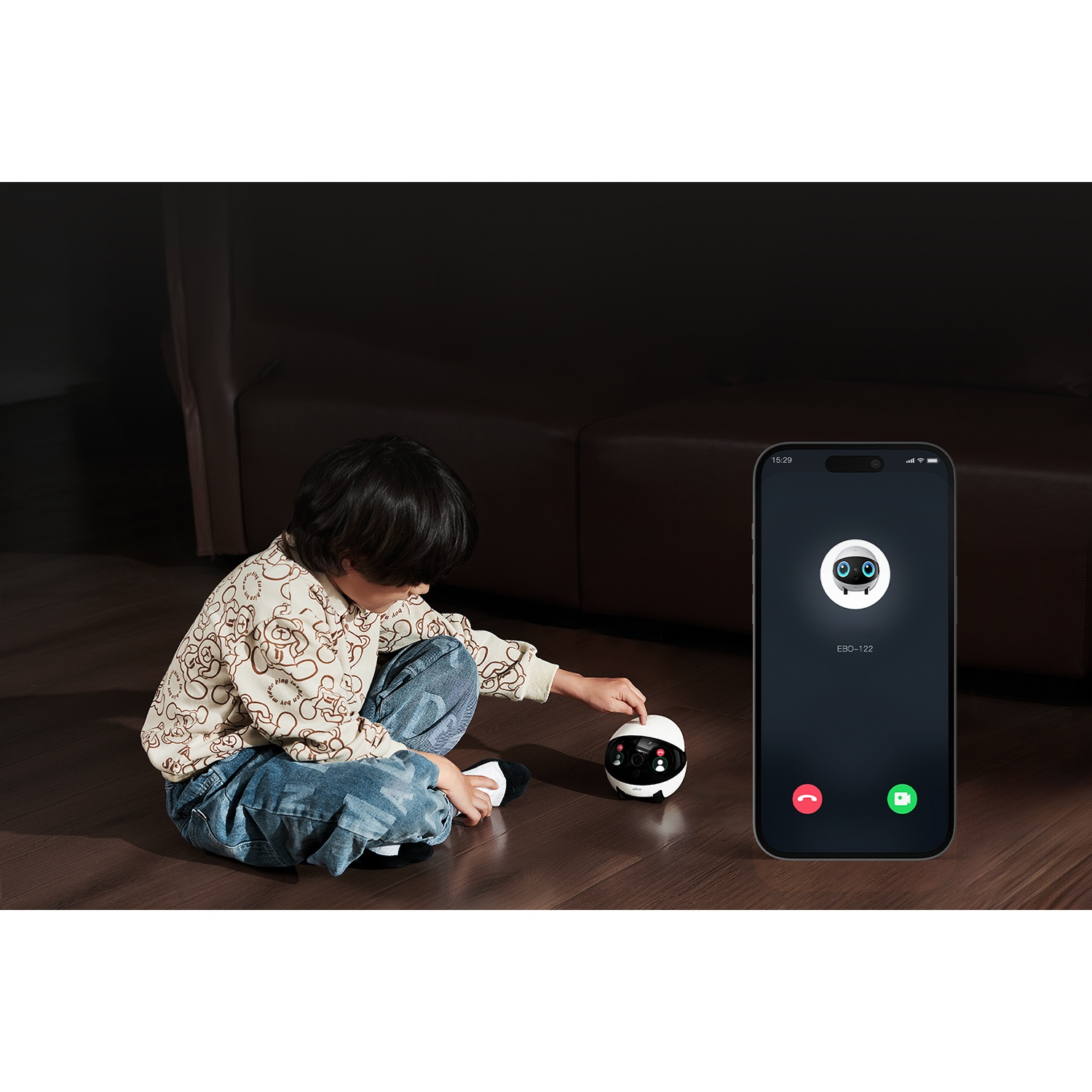 Un bambino interagisce con un giocattolo robot su un pavimento in legno. Un telefono mostra una videochiamata con il robot. Divano scuro sullo sfondo.