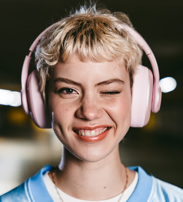 Una donna con capelli biondi corti fa l'occhiolino, indossa cuffie rosa e una camicia blu. Sorride ampiamente.