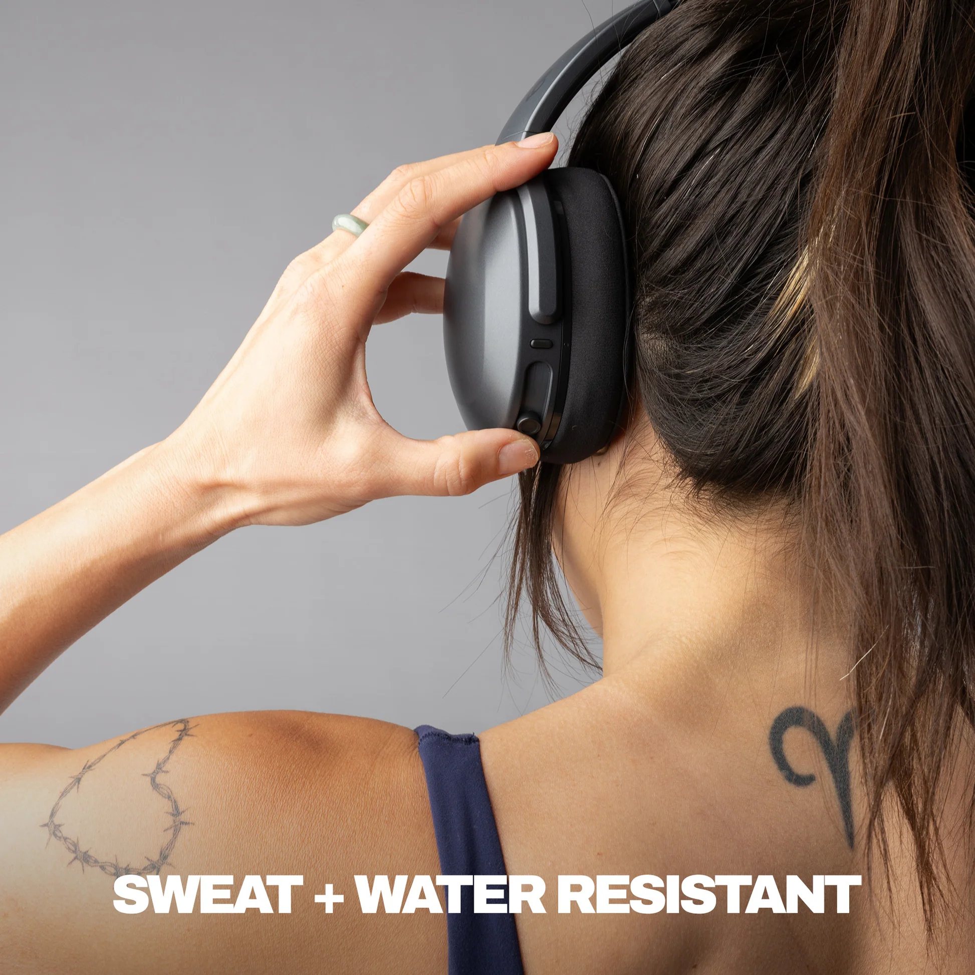 Una donna con le cuffie, mano sul dispositivo. Tatuaggio sulla spalla. Testo: 'SWEAT + WATER RESISTANT'.