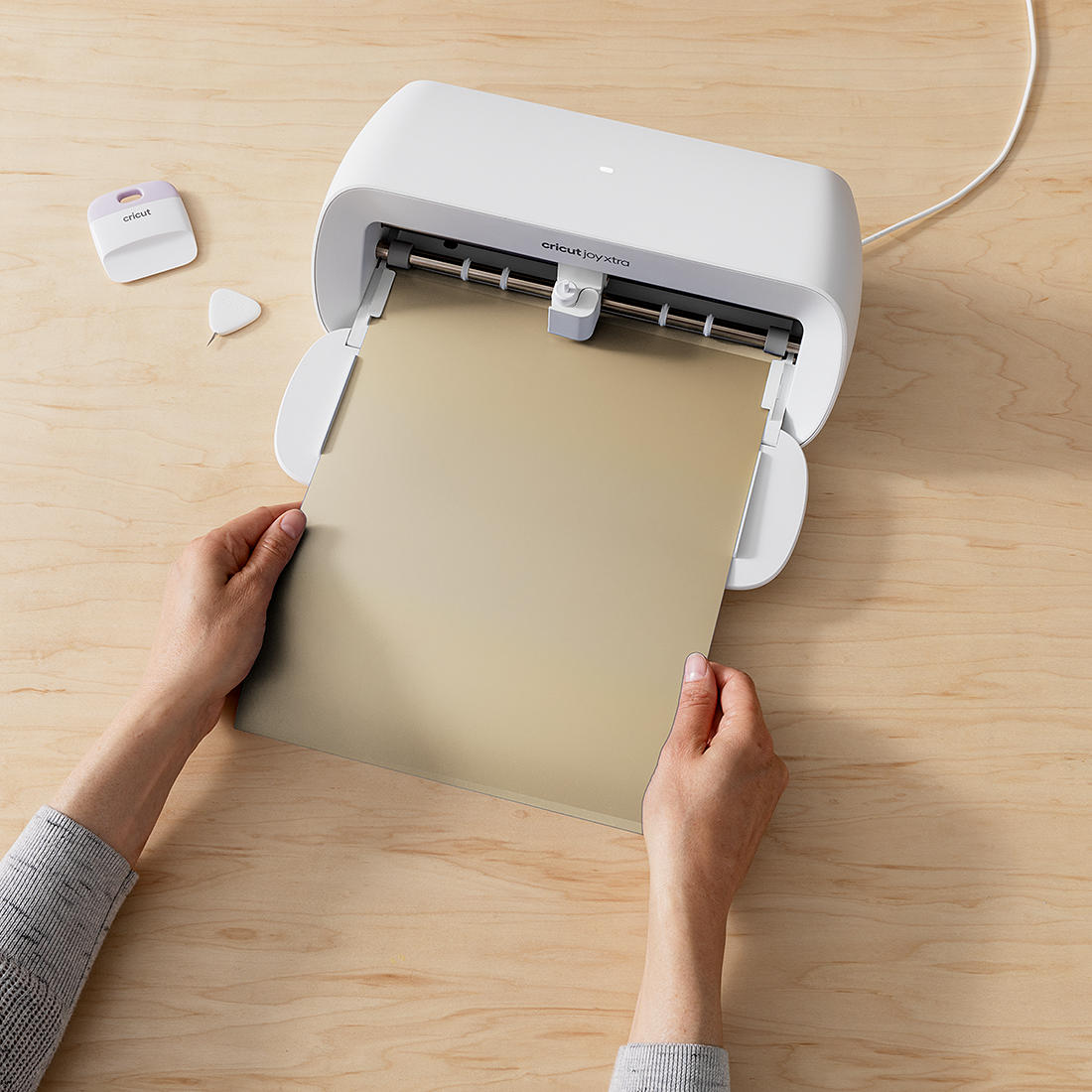 Hände halten ein goldenes Blatt in einer weißen Cricut Joy Xtra Maschine. Ein lila Schaber und ein weißes Werkzeug liegen auf dem Tisch.