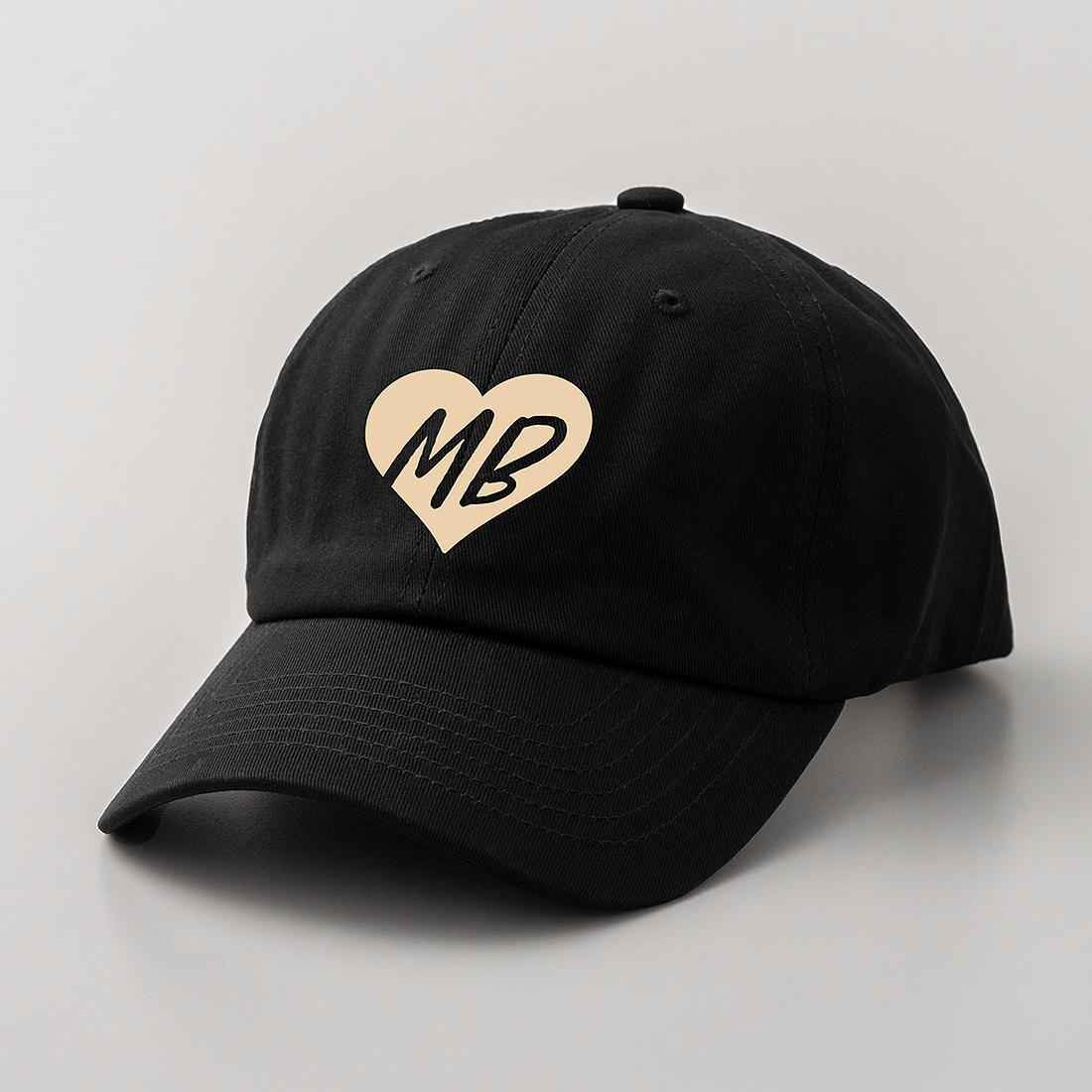 Eine schwarze Kappe mit einem beigeherzigen Logo. Das Logo zeigt die Buchstaben 'MB'. Die Kappe befindet sich auf einer grauen Oberfläche.