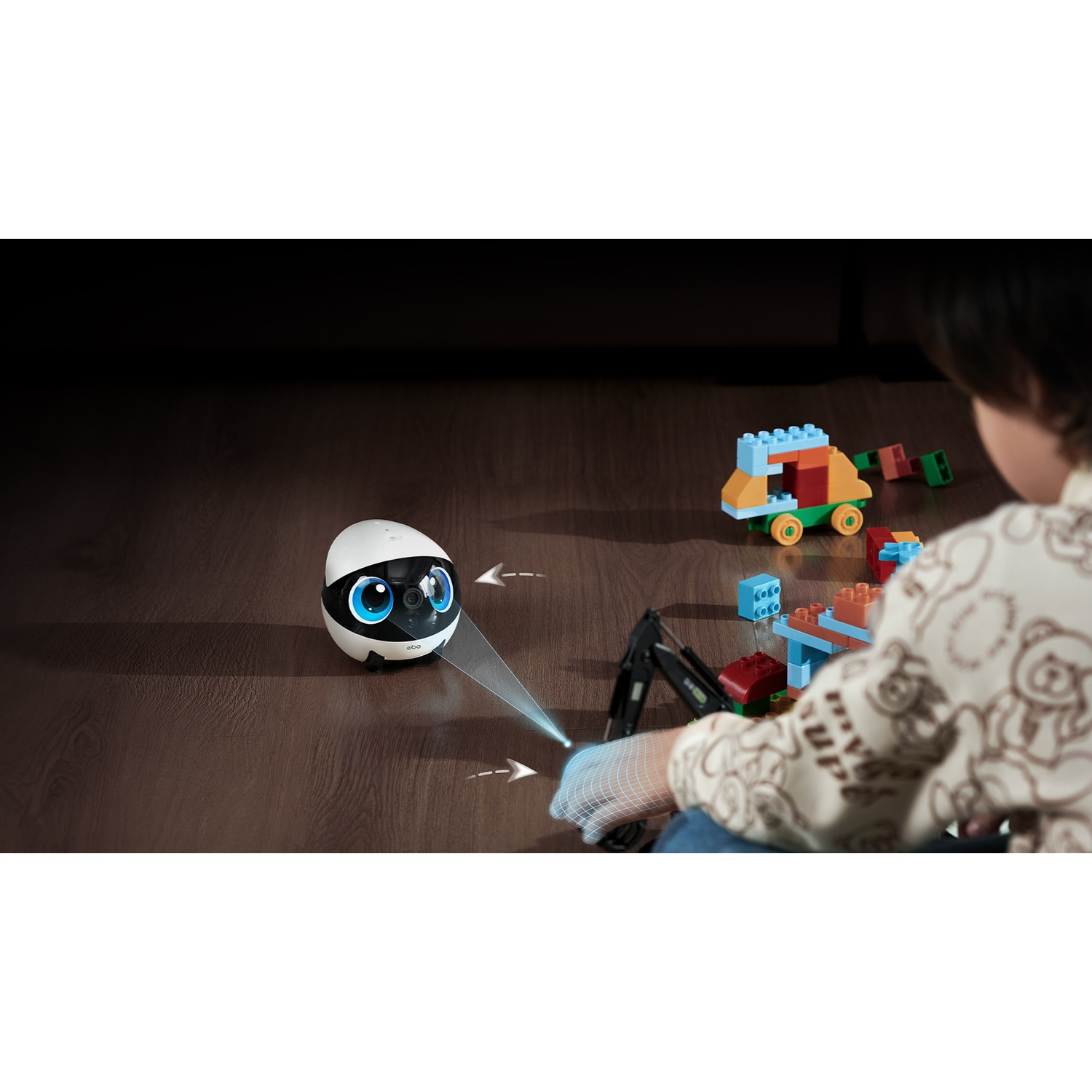 Un robot bianco con grandi occhi blu è sul pavimento. Un bambino gioca con i mattoncini. Sono visibili blocchi e un camion giocattolo.