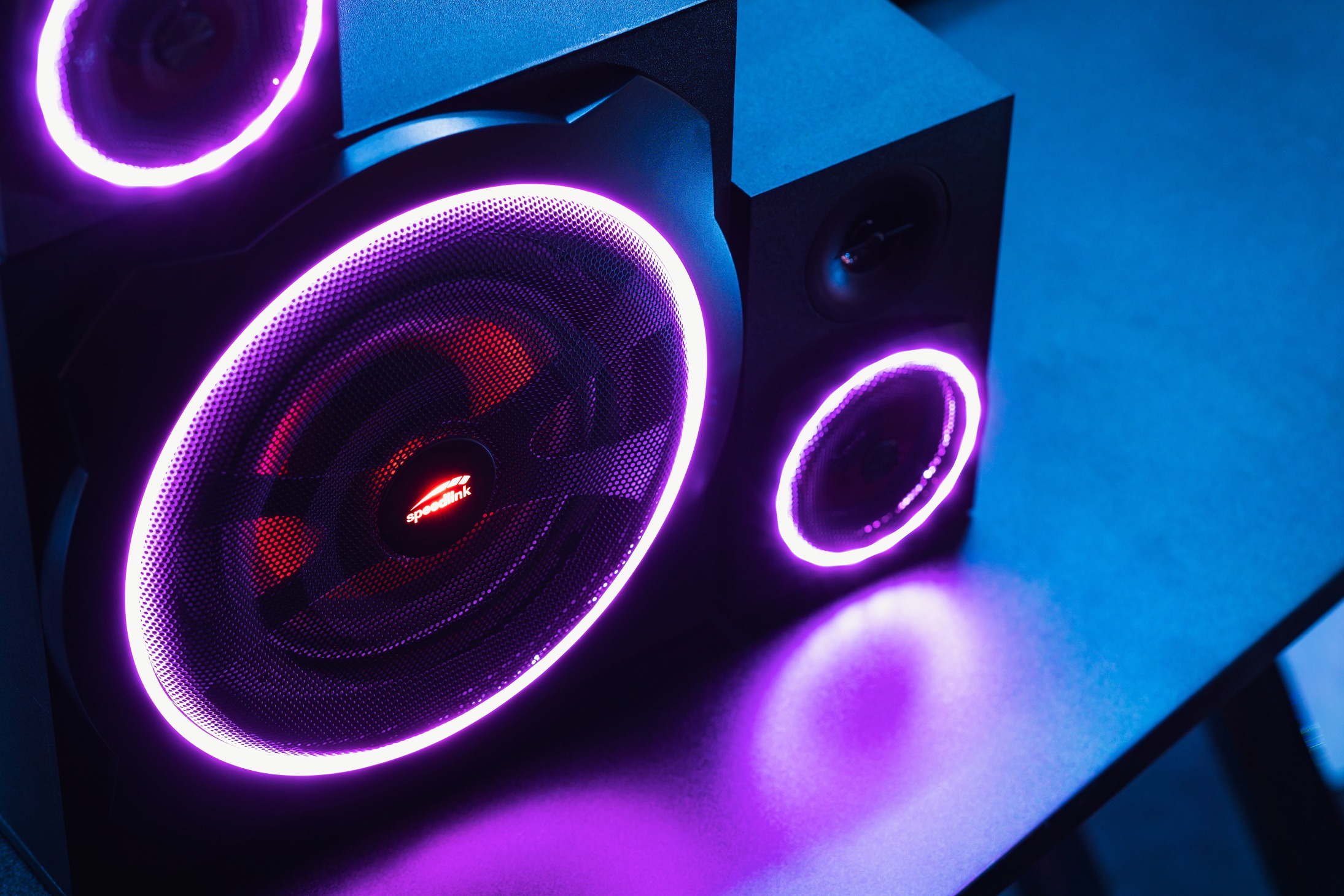 Ein schwarzes Lautsprechersystem mit violetten Neonlichtern. Die Lautsprecher haben ein rotes Gitter und das Speedlink-Logo ist sichtbar.