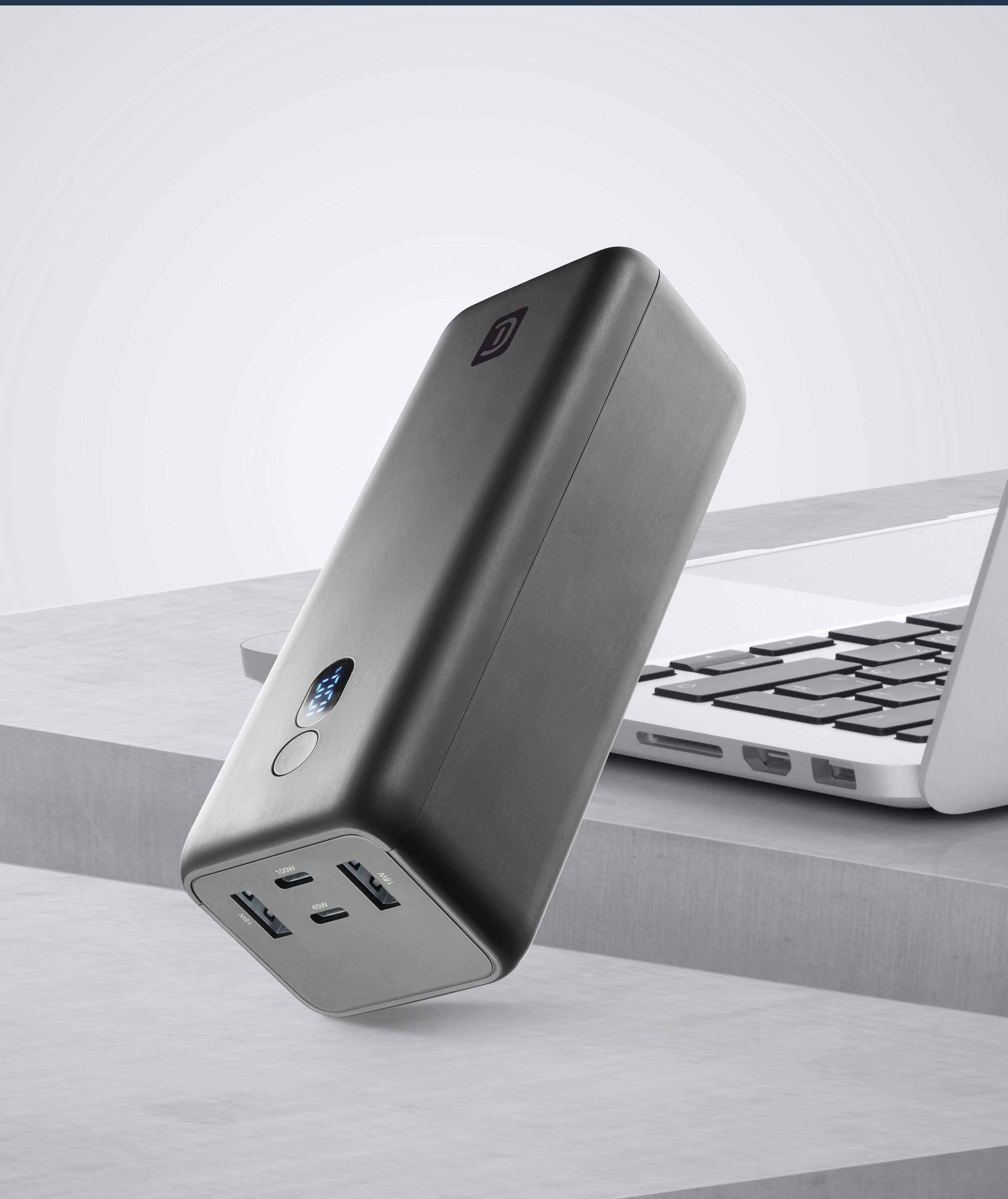 Siyah bir powerbank, bir dizüstü bilgisayarın yanında gri bir yüzey üzerinde duruyor. Powerbank'in dijital bir ekranı var.