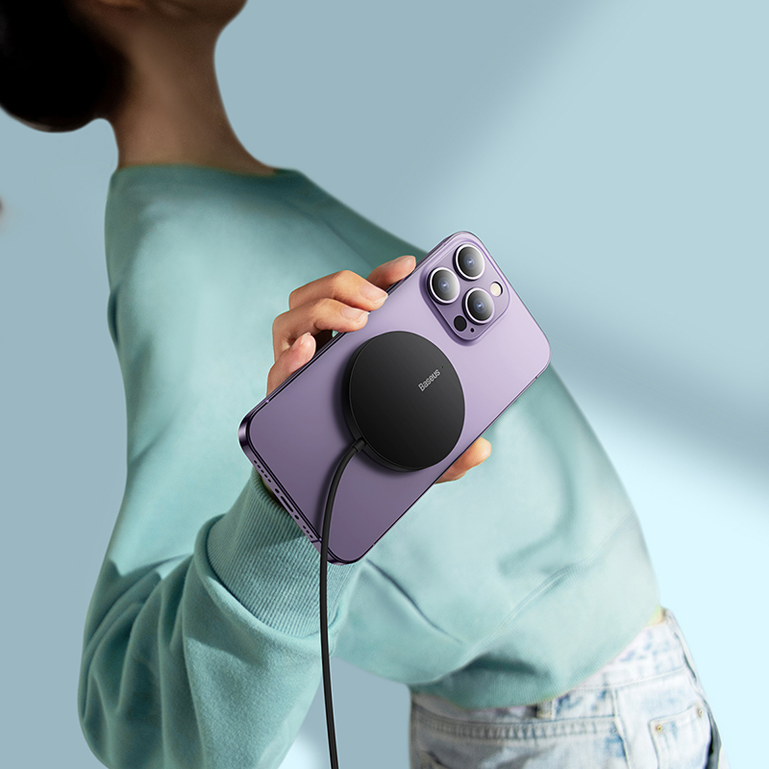 Una persona sostiene un teléfono morado con un cargador negro. Viste un suéter verde azulado. El fondo es azul claro.