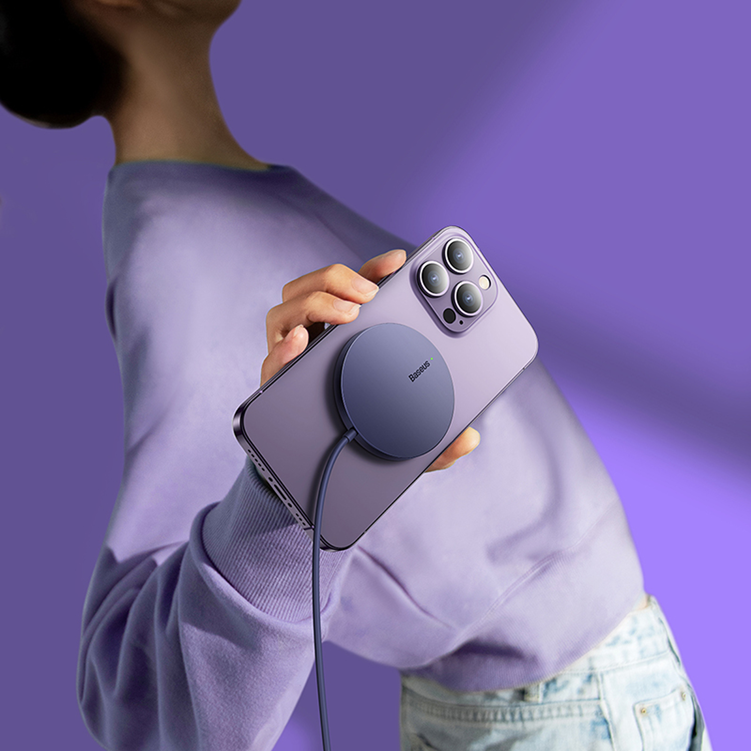 Una persona sostiene un teléfono morado con un cargador magnético. El fondo es un degradado de morado. La persona lleva un suéter morado.