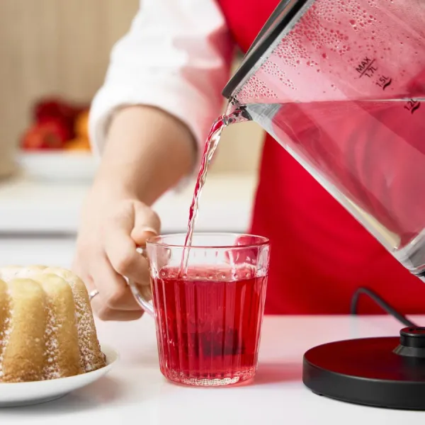 Osoba wlewa czerwony płyn ze szklanego czajnika do przezroczystego szklanego kubka. Ciasto i owoce są w tle.