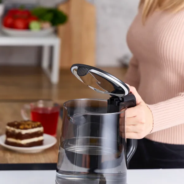 Kobieta otwiera czajnik elektryczny. Ciasto i szklanka czerwonego płynu na stole. Pomidory i zieleń w tle.