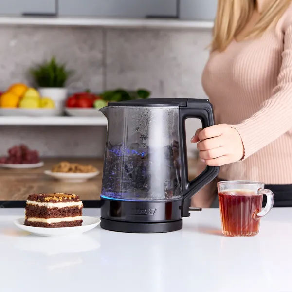 Kobieta trzyma czarny czajnik z wrzącą wodą. Na stole filiżanka herbaty i ciasto. Owoce w tle.