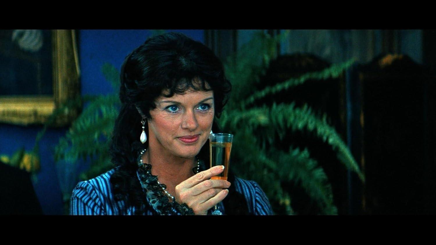 Eine Frau mit dunklem Haar hält ein Getränk. Sie trägt Ohrringe und ein blau gestreiftes Oberteil. Im Hintergrund sind eine Pflanze und ein Gemälde zu sehen.