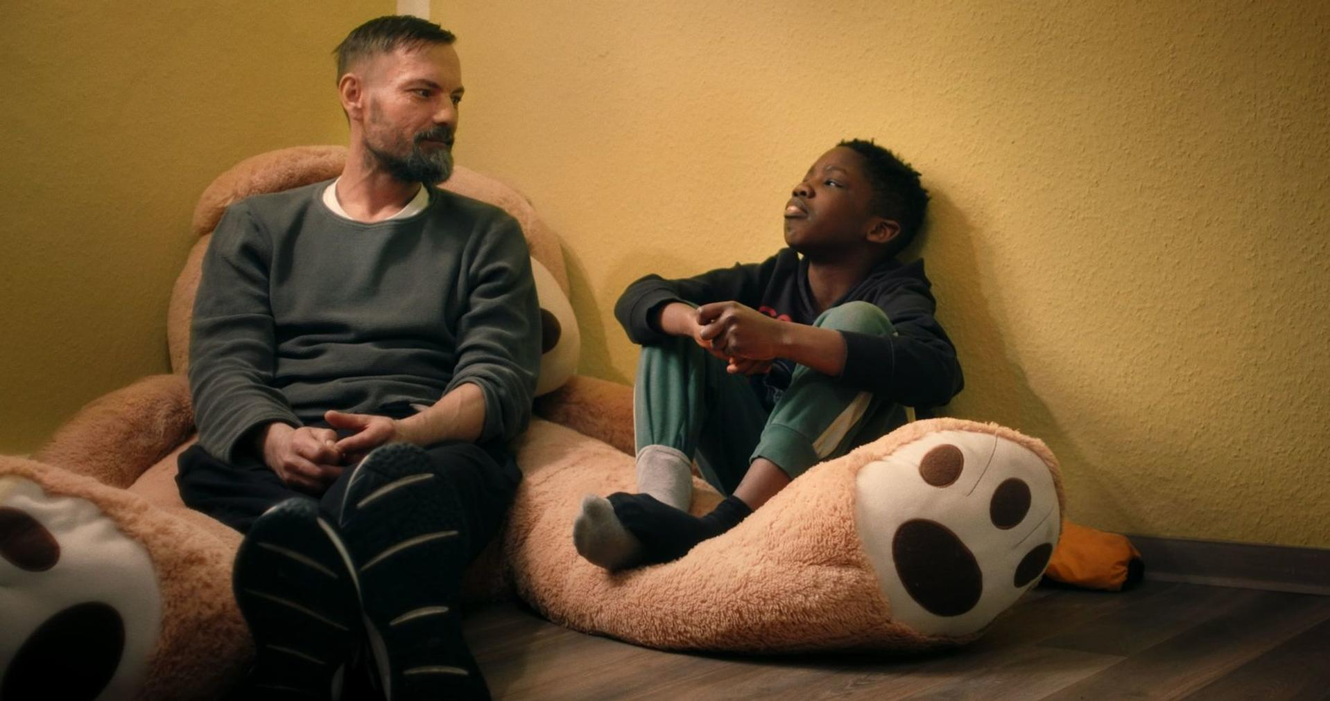 Ein Mann und ein Junge sitzen auf einem großen Teddybären. Der Mann blickt den Jungen an, der nach oben schaut. Gelbe Wand im Hintergrund.