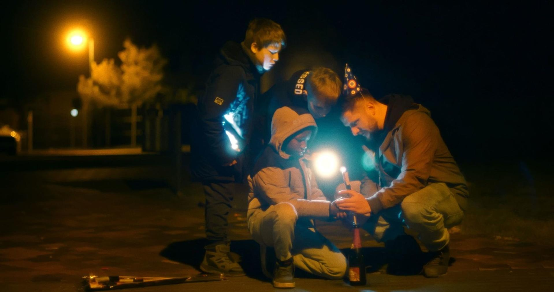 Vier Personen bei Nacht mit einem brennenden Wunderkerze. Einer kniet. Eine Flasche ist im Vordergrund, eine Straßenlaterne dahinter.