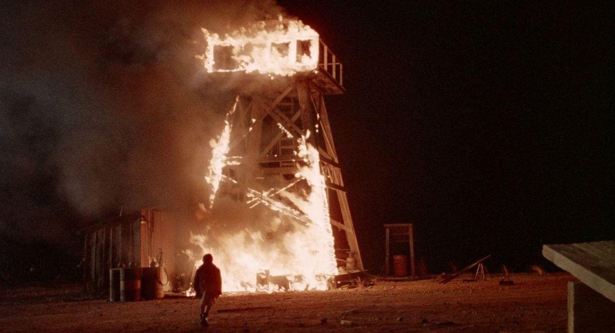 Ein hölzerner Wachturm brennt nachts. Eine Person geht vom Feuer weg. Fässer und ein kleines Gebäude sind links.