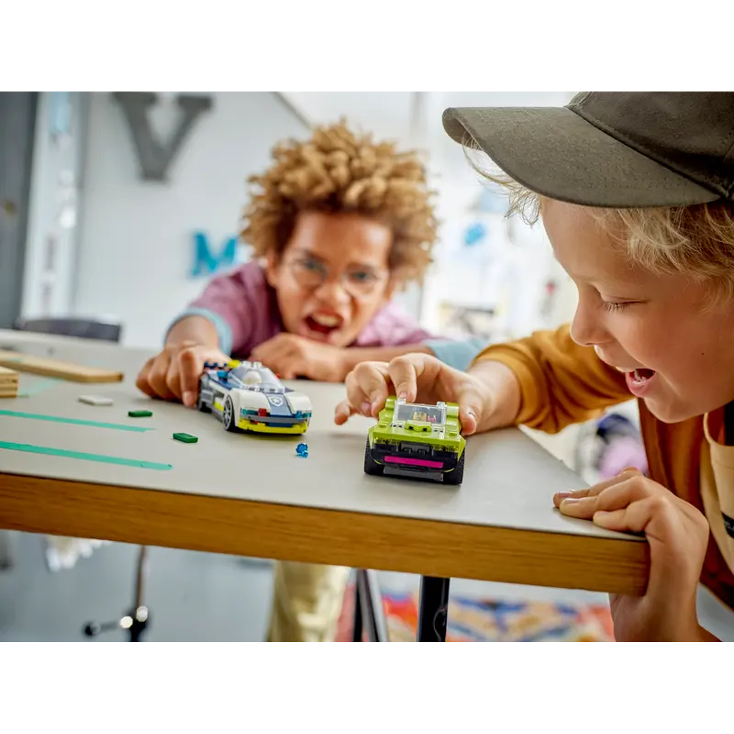 Two boys play with toy cars on a table. One boy wears a cap. The cars are blue and green. The background has a letter M.