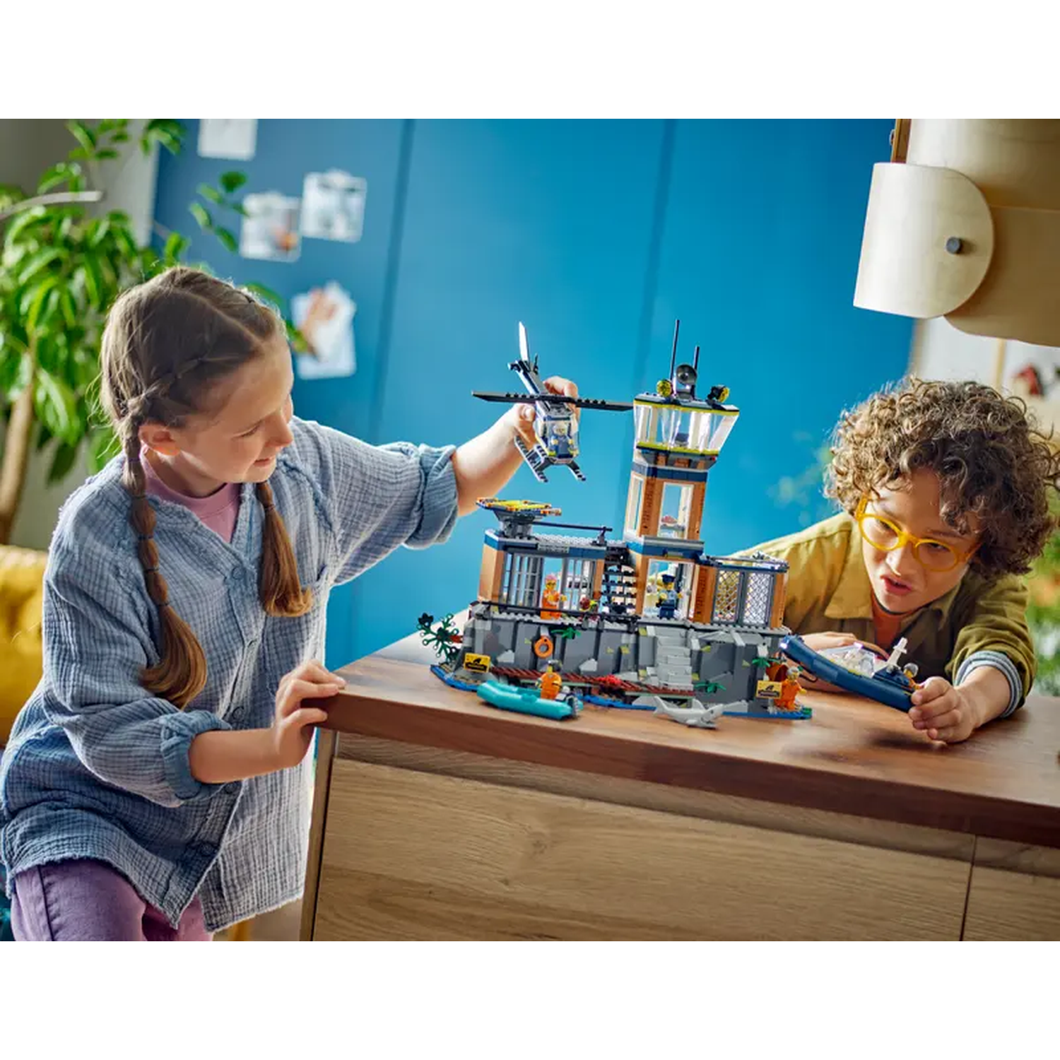 Two kids play with a Lego set. A girl holds a helicopter, a boy with glasses plays with a boat. The set is a prison.