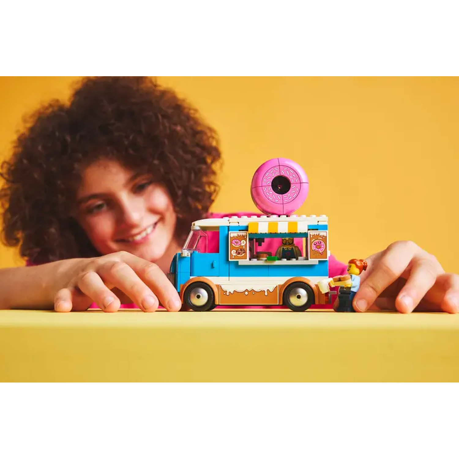 A girl smiles at a toy food truck. The truck is blue and white with a pink donut on top. A Lego figure is beside it.