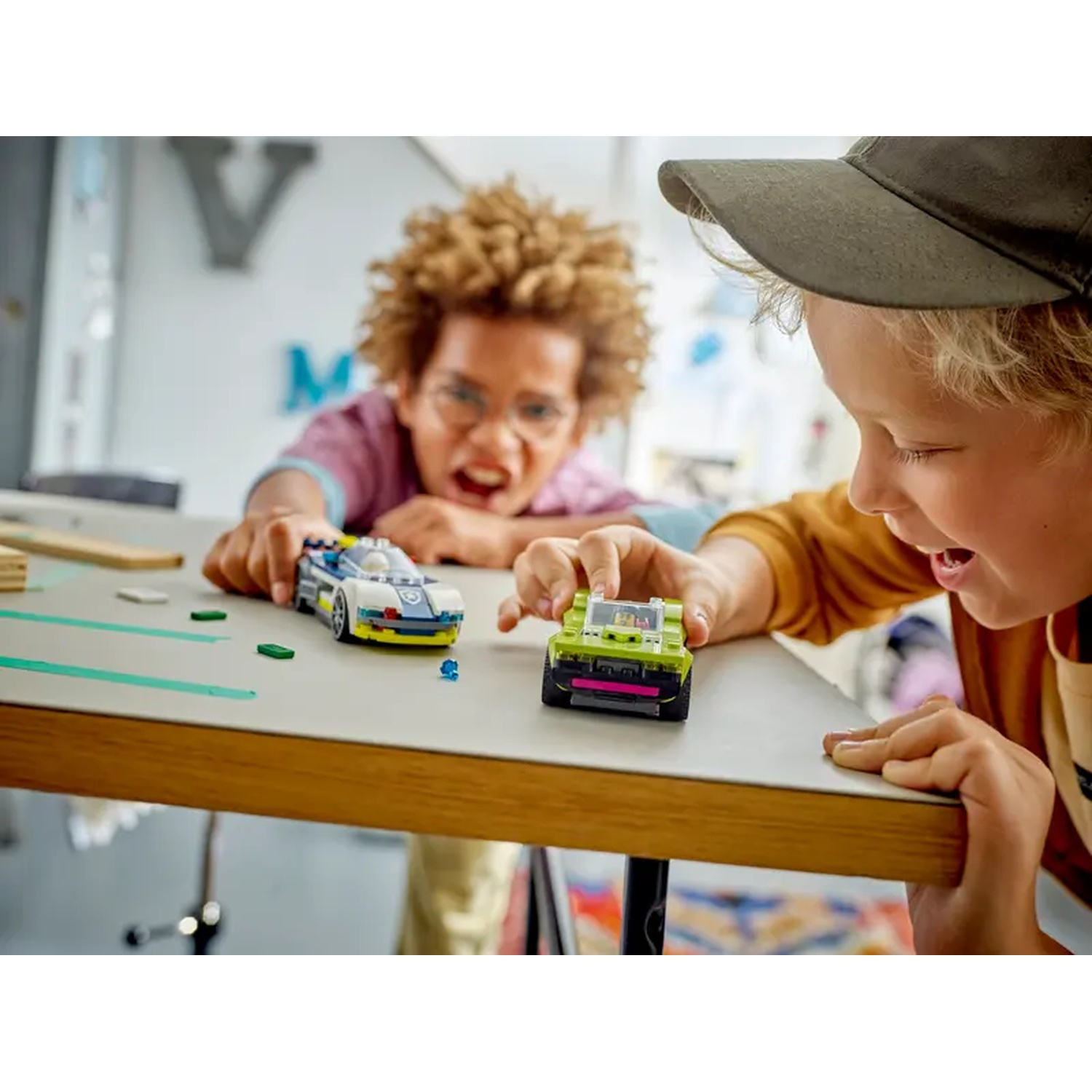 Two children play with toy cars on a table. One child wears a cap, the other has curly hair. Cars are green and white.
