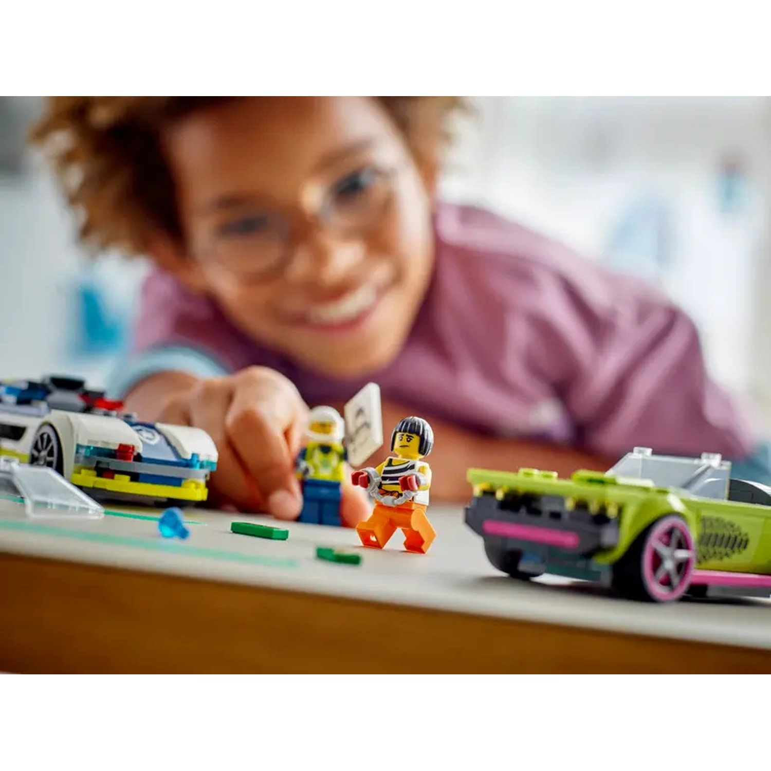 A smiling child plays with Lego cars and figures. The cars are various colors. The child is holding a figure.