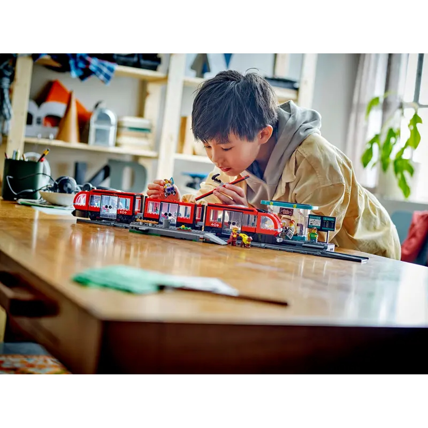 A boy is playing with a toy train set on a wooden table. The train is red, the boy is wearing a yellow jacket.