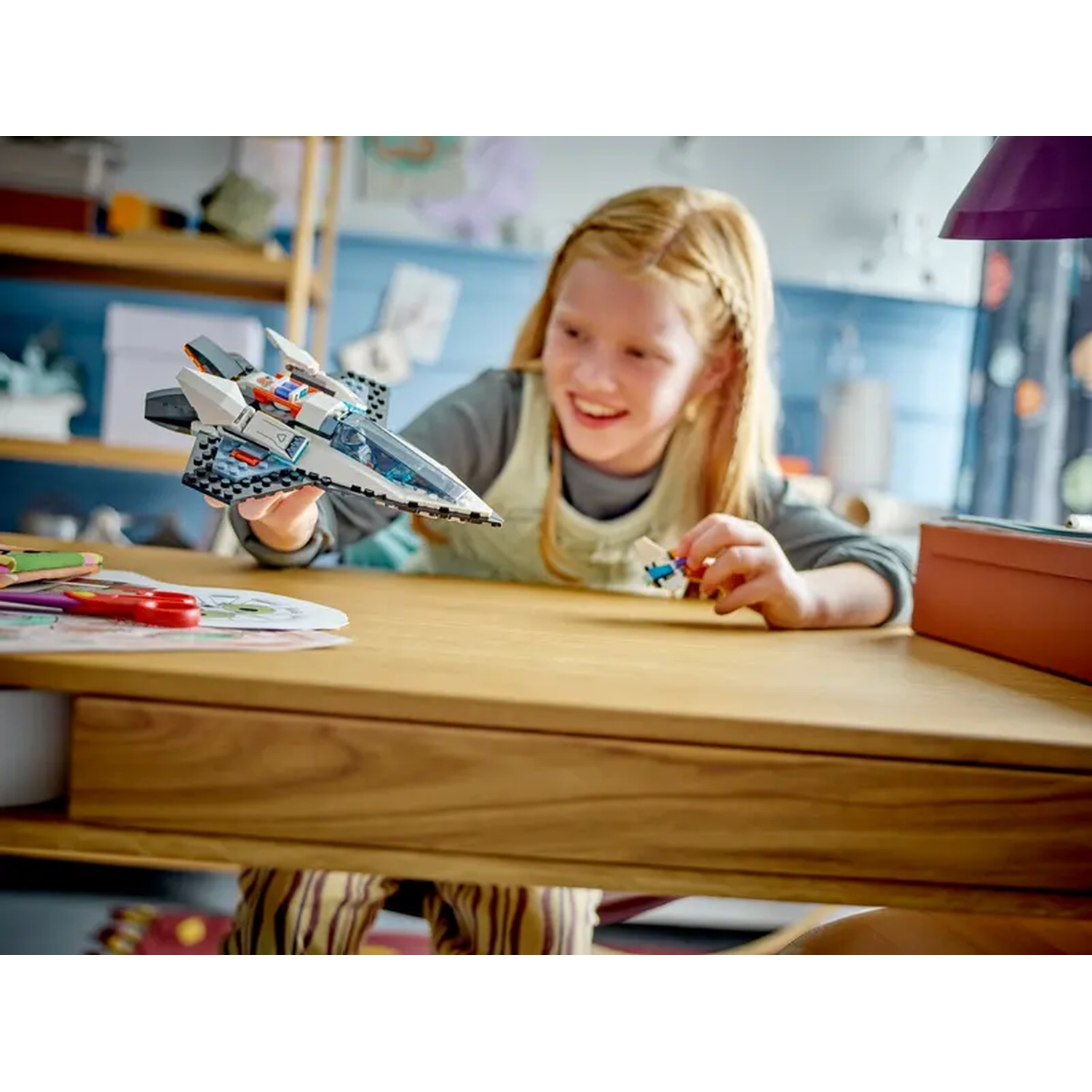 Een meisje met blond haar glimlacht terwijl ze een speelgoed ruimteschip vasthoudt. Een houten tafel en een lamp zijn in de scène.