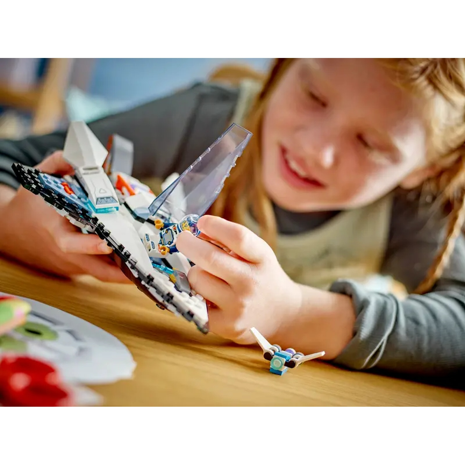 Een meisje lacht, met een Lego ruimteschip in haar handen. Een klein ruimteschip ligt op de tafel. De tafel is bruin. Haar gezicht en handen zijn scherp.