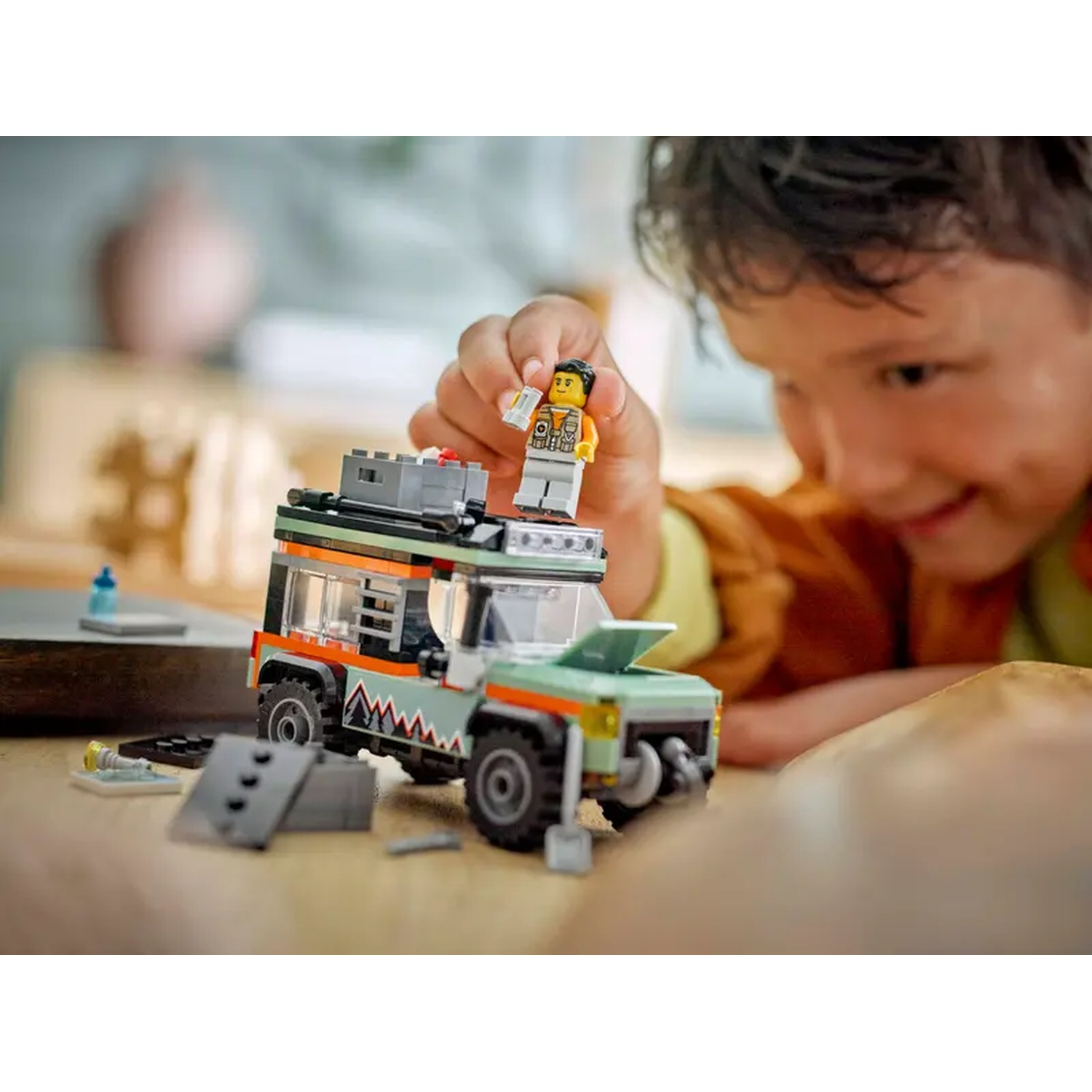 A child is playing with a Lego car. The car is green and orange. A Lego figure is on top. The child is smiling.
