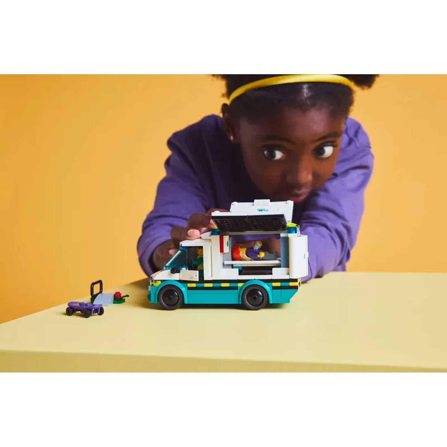 Een meisje kijkt naar een Lego-ambulance. De bus staat op een gele tafel. Een klein karretje links. Gele achtergrond.