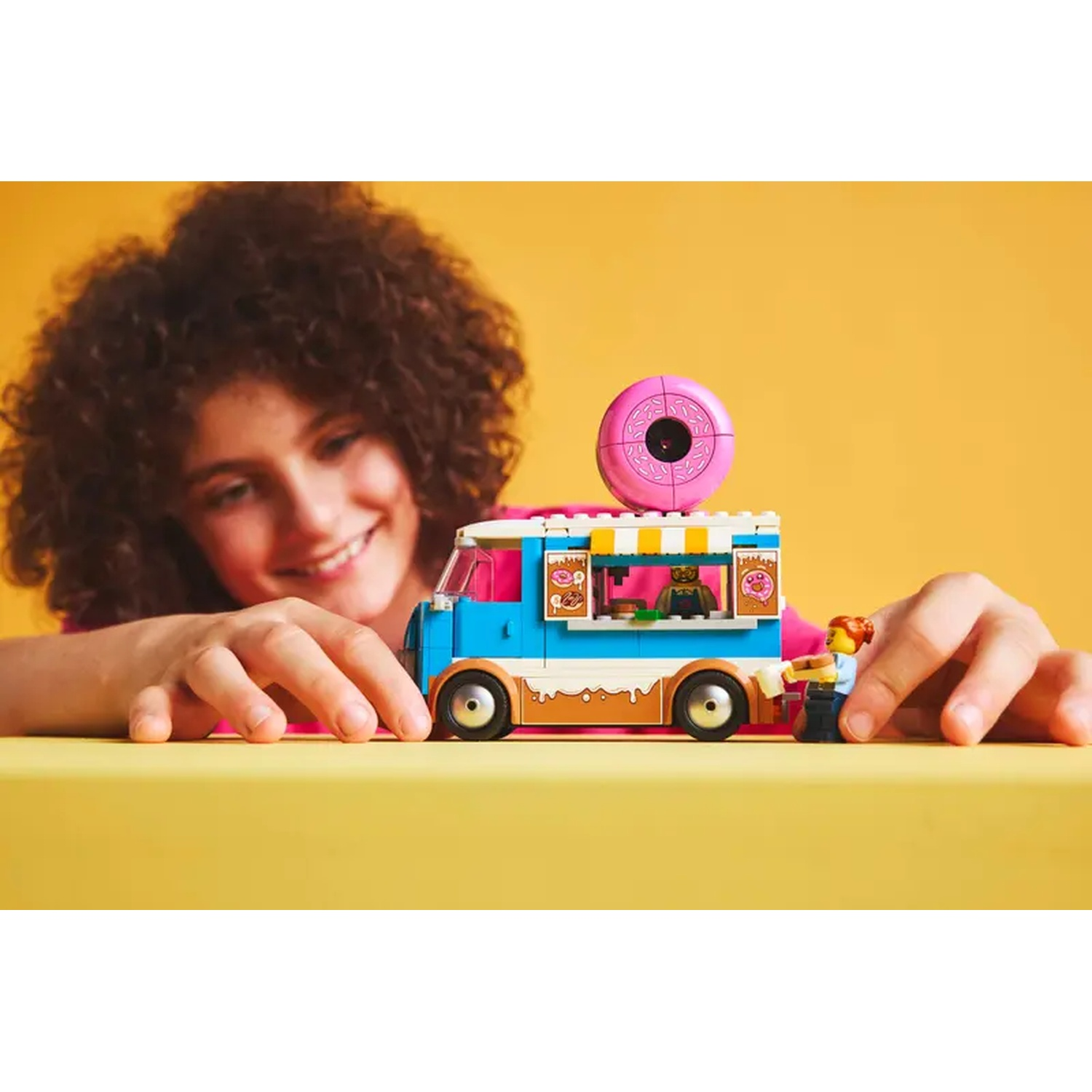 Een meisje glimlacht naar een speelgoed donut truck. De truck is blauw, roze en bruin. Een klein figuurtje staat bij de truck.