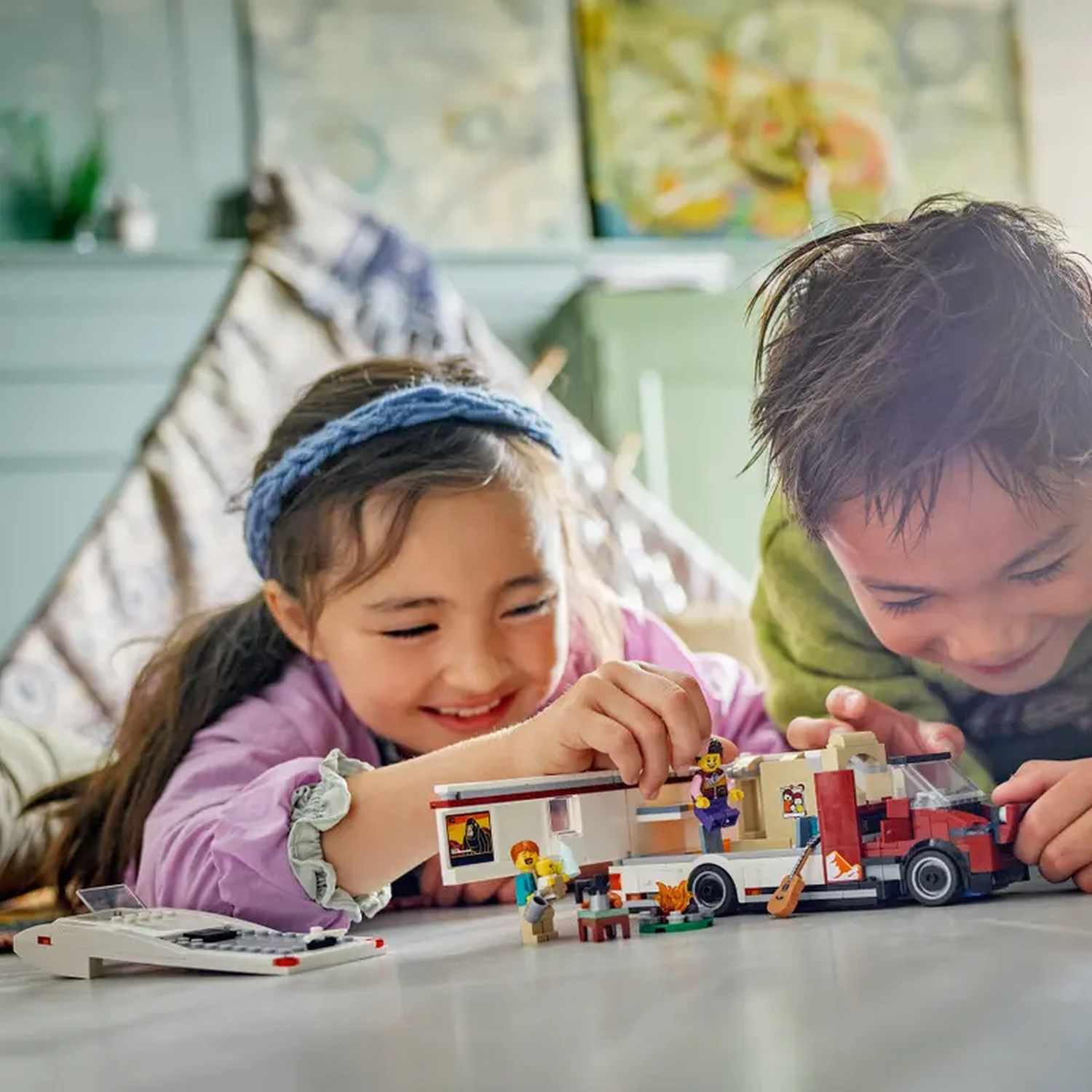 Two children playing with a Lego camper van. The girl is wearing a blue headband and the boy a green shirt. A Lego plane is on the left.