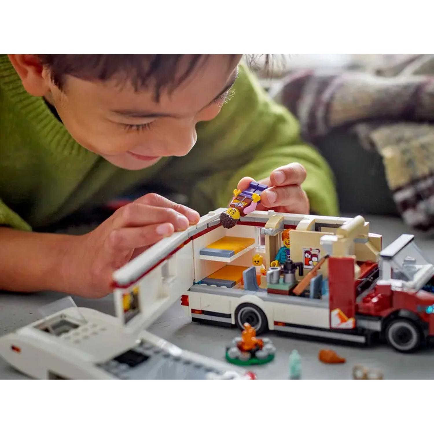A boy playing with a Lego camper van. The van has an open door, revealing interior details and mini-figures. A small campfire is in front.