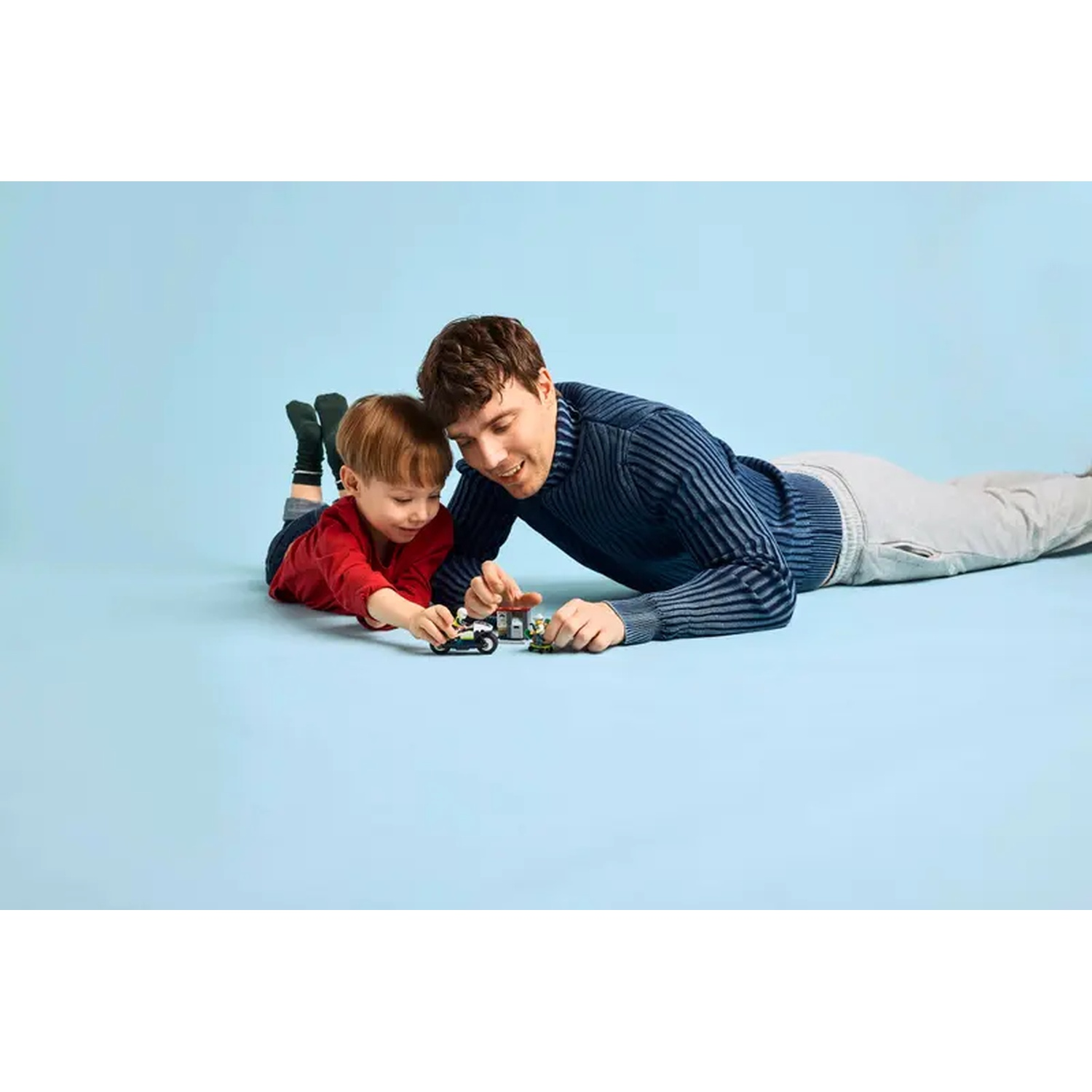 A man and a child are lying on the floor. They are playing with toy vehicles. The background is light blue.