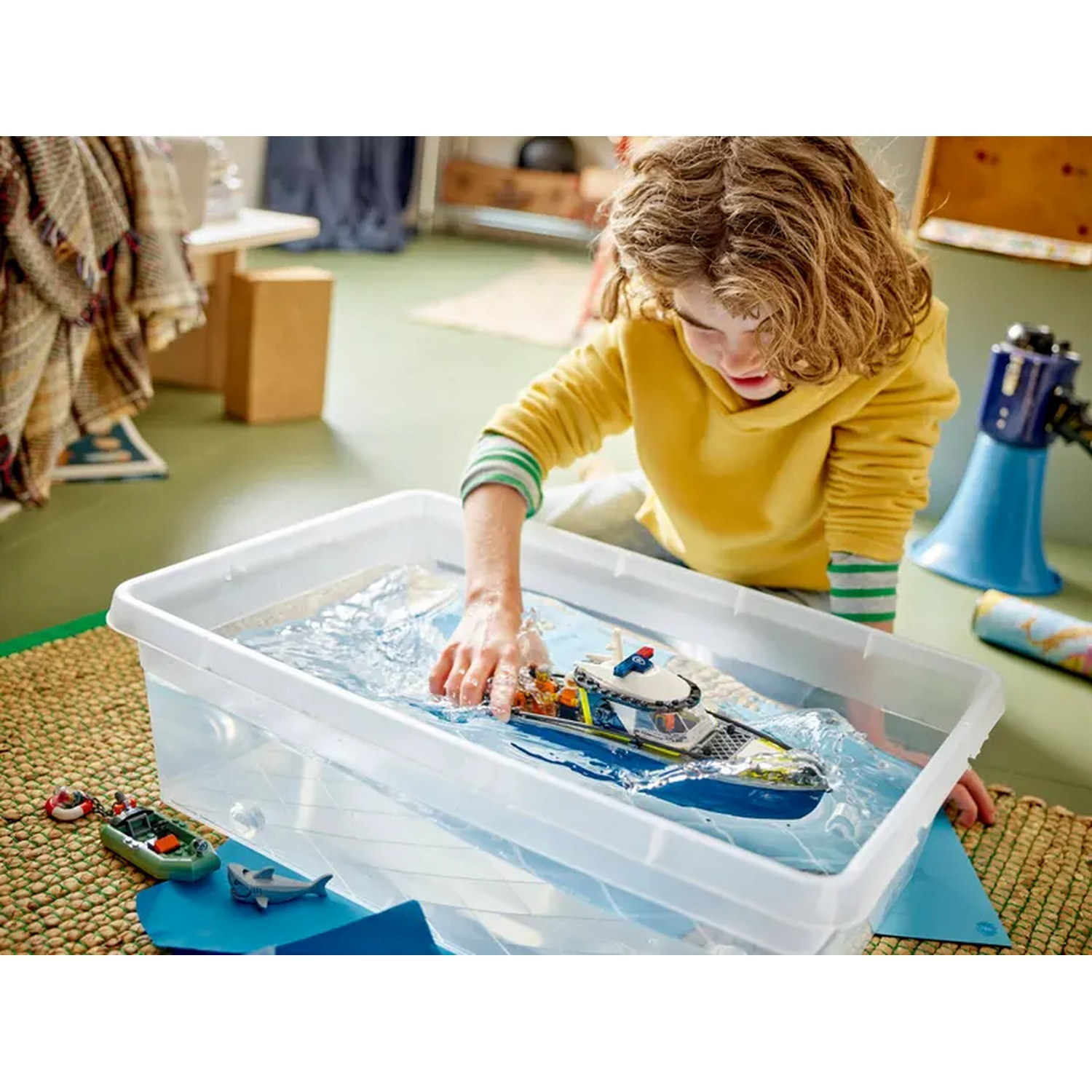 Een kind speelt met een speelgoedboot in een bak gevuld met water. Andere speeltjes, waaronder een kleine boot en een haai, liggen in de buurt op een mat.