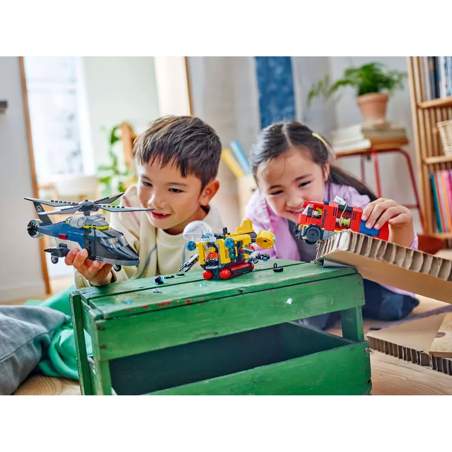Two children playing with toy vehicles. A boy holds a helicopter, a girl a fire truck. They're near a green box and cardboard.