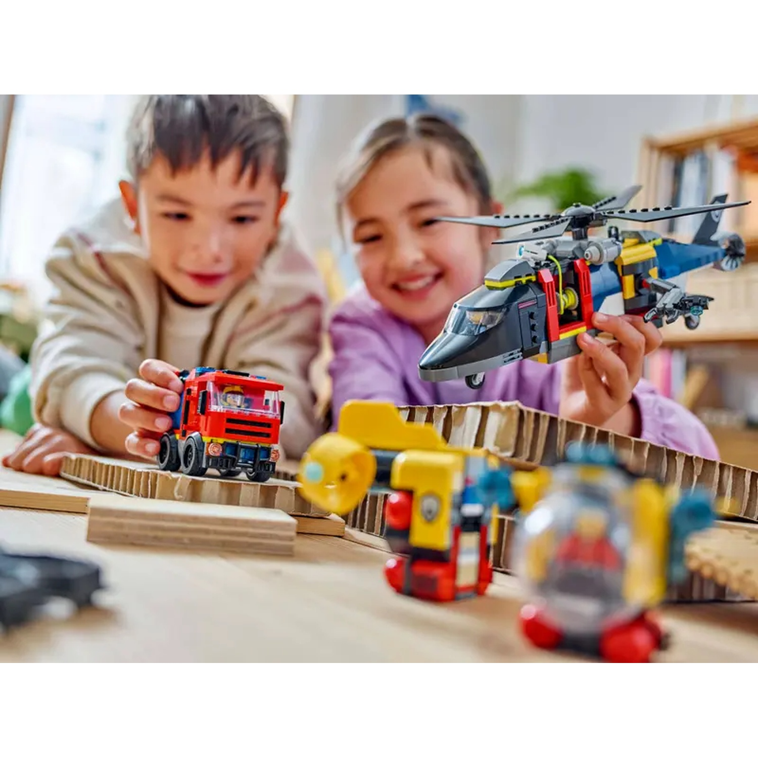 Two children playing with toy vehicles. A red fire truck, a yellow submarine, and a black and blue helicopter are visible.