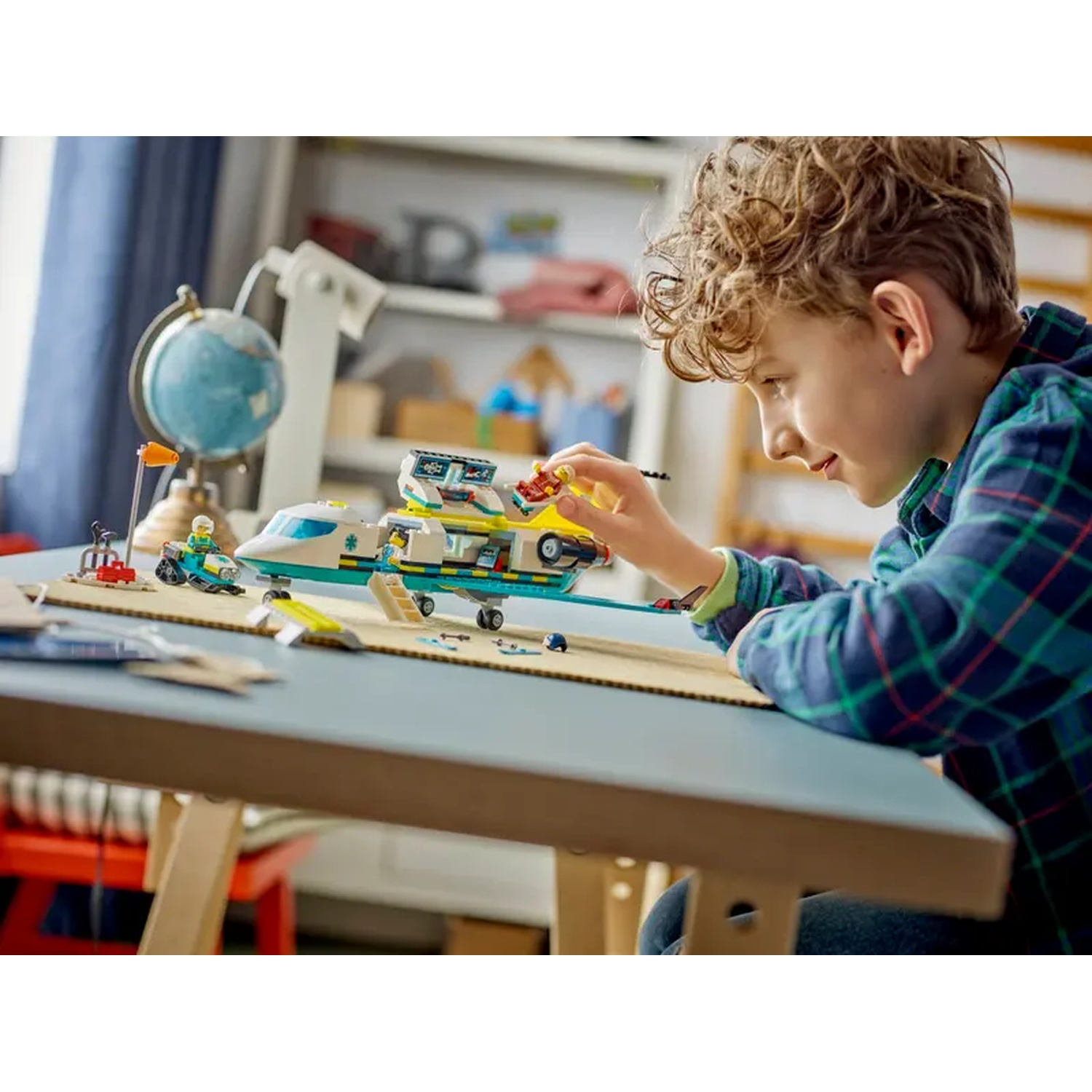 Een jongen speelt met een Lego-vliegtuigset. Hij is gefocust op het speelgoed. Een wereldbol en een plank staan op de achtergrond.