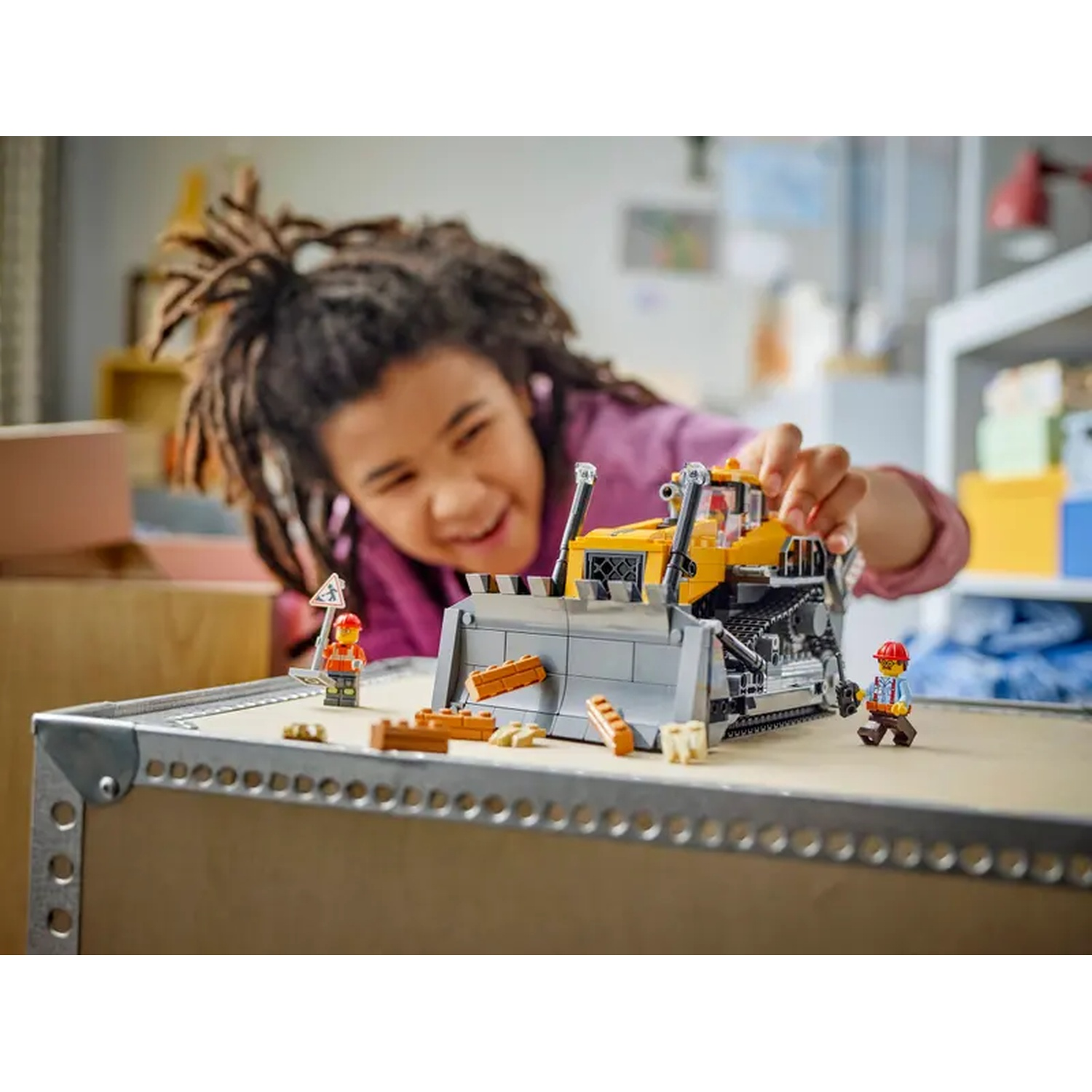 A child plays with a yellow toy bulldozer. Lego figures and construction blocks are on a table. The child smiles.