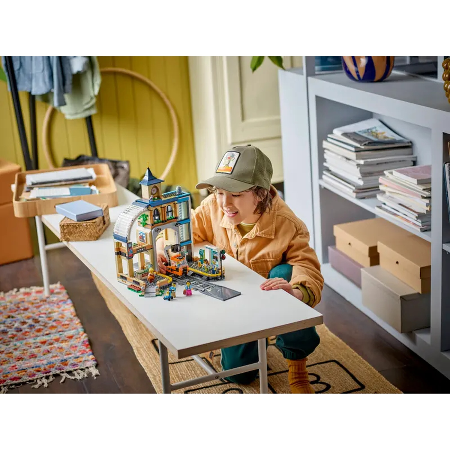 Een kind speelt met een Lego-set op een witte tafel. De set toont een gebouw en een trein. Het kind draagt een hoed.